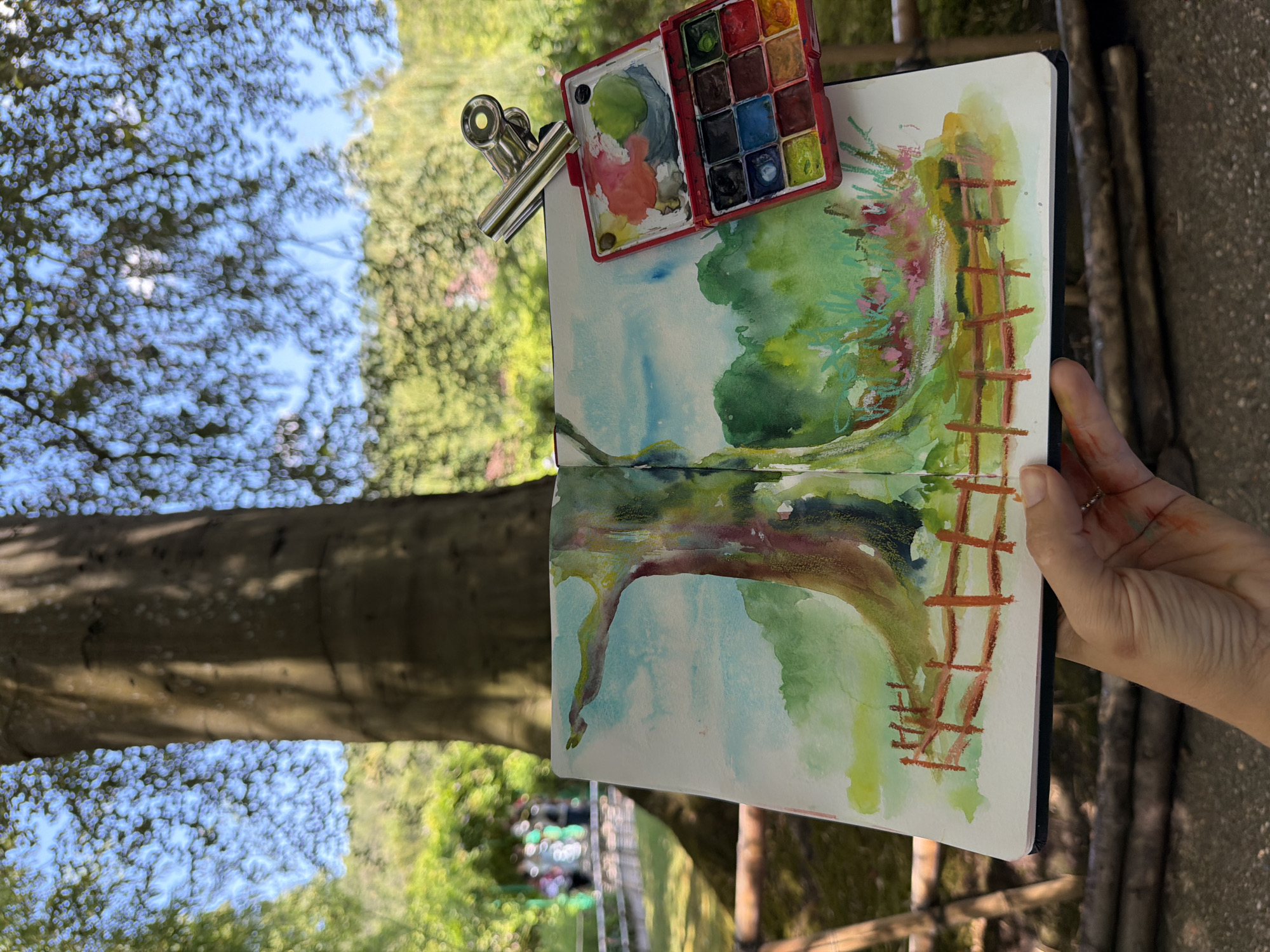 An artist is sketching a watercolor painting of a tree and garden in Giverny, France. The painting is open in a sketchbook, held by the artist's left hand, and shows a large tree trunk on the left, with a fence in the foreground. A watercolor palette is clipped to the top of the sketchbook. In the background, a garden with blooming flowers and other trees is visible. A few people are seen walking in the garden in the distance. The scene is set outdoors, with dappled sunlight filtering through the trees.
