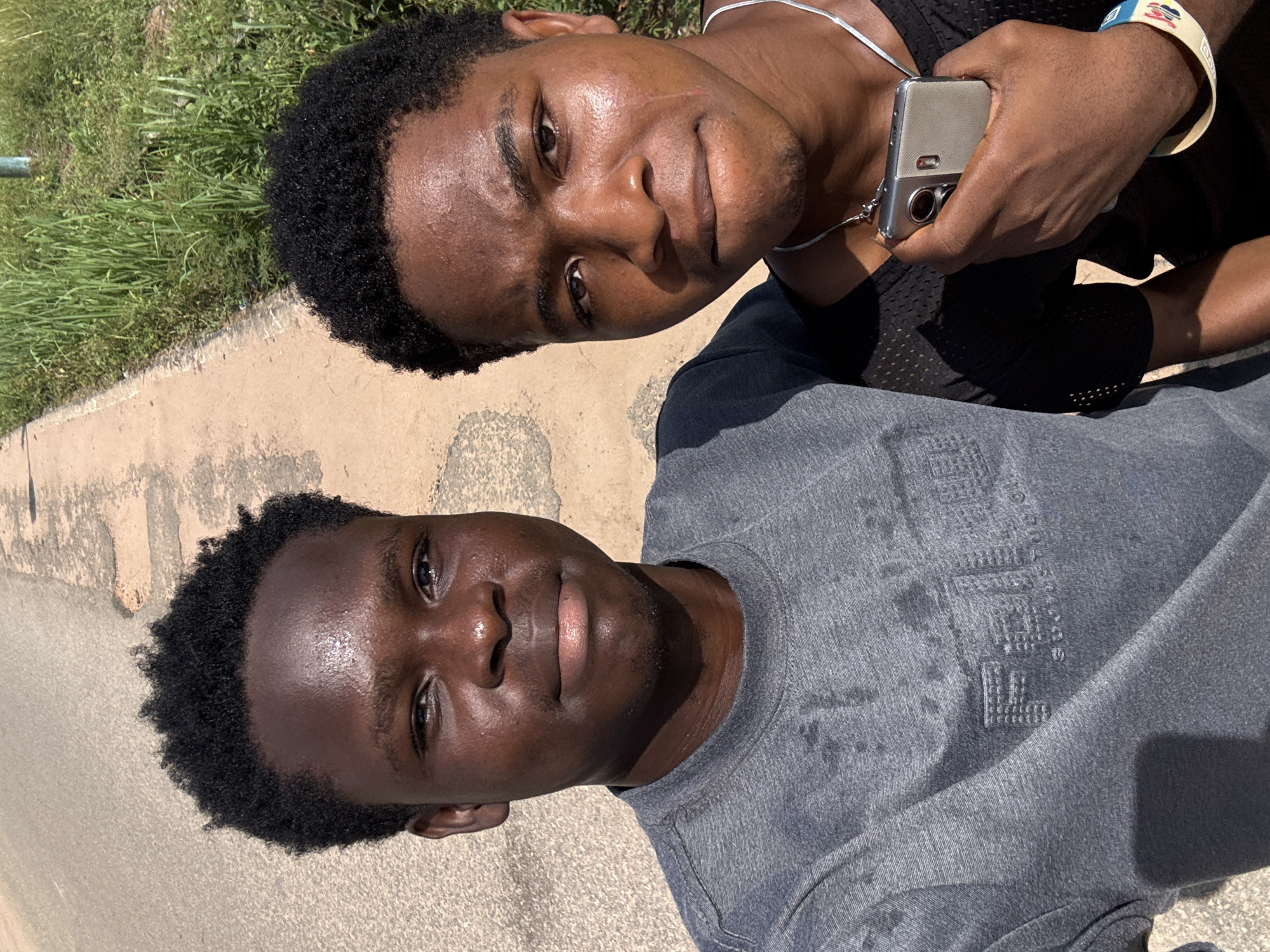 Two young men are captured in a bright outdoor photo, likely taken in Ihiagwa, Nigeria. The image is oriented vertically, showing the men positioned side-by-side, facing the camera.
The man in the foreground, on the left, has short, dark, curly hair and is wearing a grey long-sleeve shirt. His face appears slightly shiny, possibly from sweat or oil, and he maintains a neutral expression while looking directly at the camera. An embossed logo, clearly visible on his shirt, reads "TRUE RELIGION BRAND JEANS" along with its characteristic horseshoe symbol.
The man in the background, on the right, also has short, dark, curly hair. He wears a black mesh tank top and a white necklace. He holds a silver-colored smartphone in his right hand, suggesting he might be taking a selfie or a joint portrait. A multicolored wristband is visible on his left wrist, with text that appears to read "LAGOS S.I.U.N. 20XX". His face also shows a slight sheen, and he has a neutral to slightly smiling expression, with a faint scar visible near his right eyebrow.
The setting is outdoors under bright daylight, indicated by strong highlights on their skin and visible shadows. The ground beneath them is a light-colored paved or concrete surface. To the left and behind the men, a patch of vibrant green grass and some foliage is visible, completing the outdoor scene.