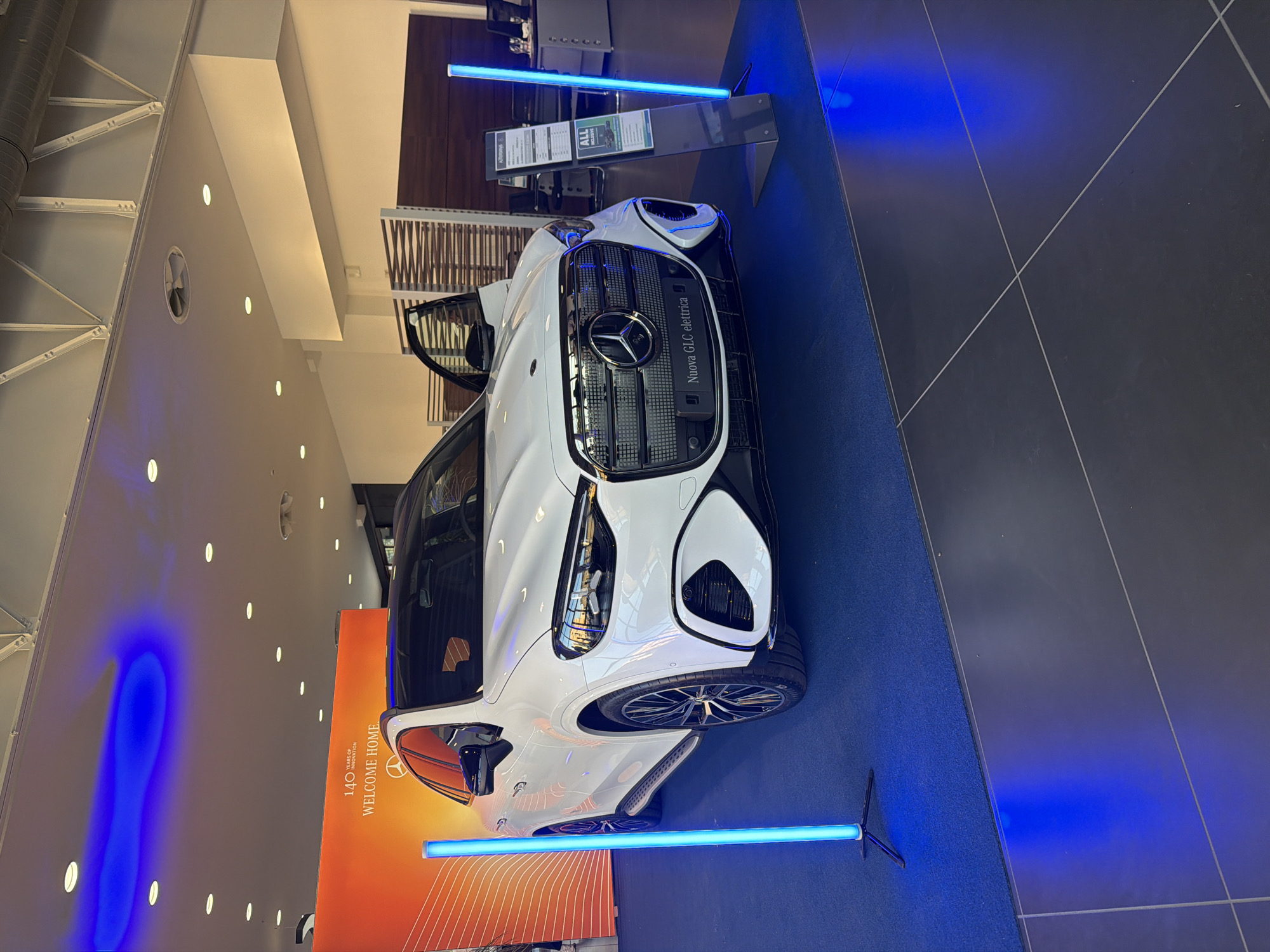 The image shows a white Mercedes-Benz SUV on display in a showroom, likely a car dealership, located in Parma, Italy. The car is positioned on a dark gray tiled floor, with a bright blue carpet area in front of it, illuminated by blue LED lights emanating from vertical light stands. Behind the car, an orange backdrop with text that appears to read "140 YEARS OF INNOVATION WELCOME HOME" is visible. The car's front doors are open, revealing part of the interior. To the right of the car, a display stand holds information brochures, including one with the text "ALL EXCLUSIVE". The overall environment is clean and well-lit, with ceiling lights and ambient blue lighting contributing to the modern showroom atmosphere.
