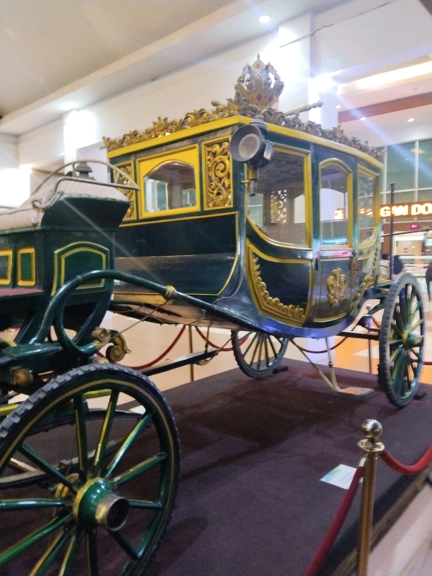 A horse-drawn carriage, ornately decorated in green and gold, is displayed indoors. The carriage features a crowned emblem on its roof, decorative scrollwork along its sides, and large spoked wheels. The carriage is positioned on a dark-colored platform, with a red velvet rope barrier in the foreground. The background shows an indoor environment with bright overhead lighting and signs, including one that reads "GAM DO....", suggesting a commercial or public space. The image does not provide information about the specific location in Desa Ngesrep, Indonesia.