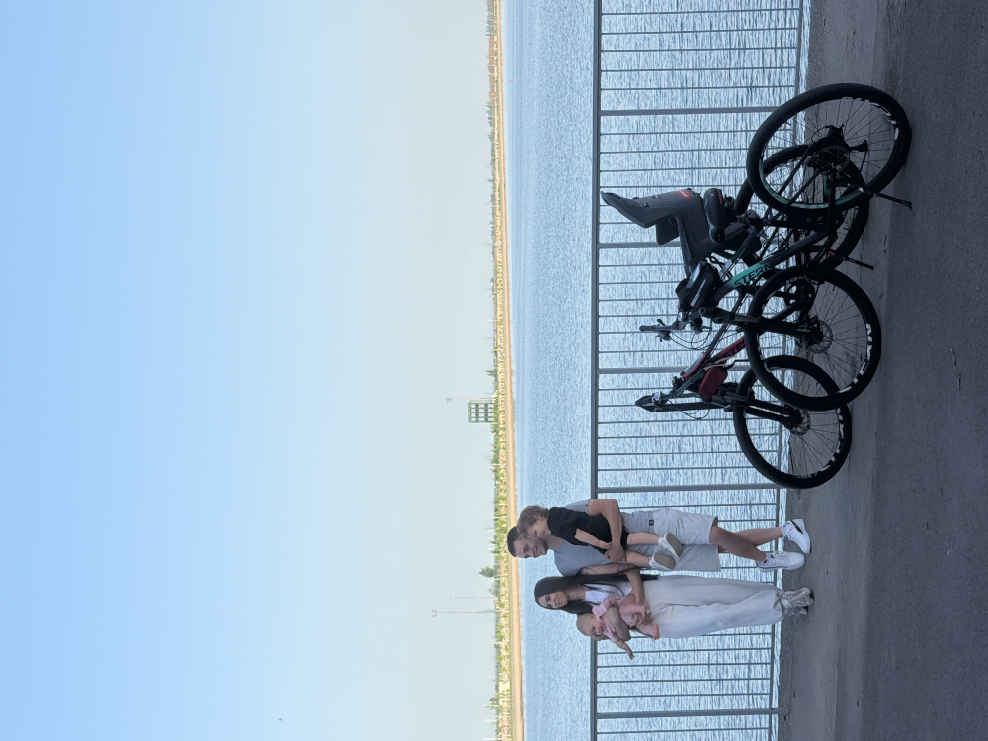 A family of four stands next to a bike rack on the embankment in Rostov-on-Don, Russia. A woman with long dark hair holds a baby, while a man holds a young child. Three bicycles are leaning against the railing. The embankment overlooks the Don River, and the sky is clear.