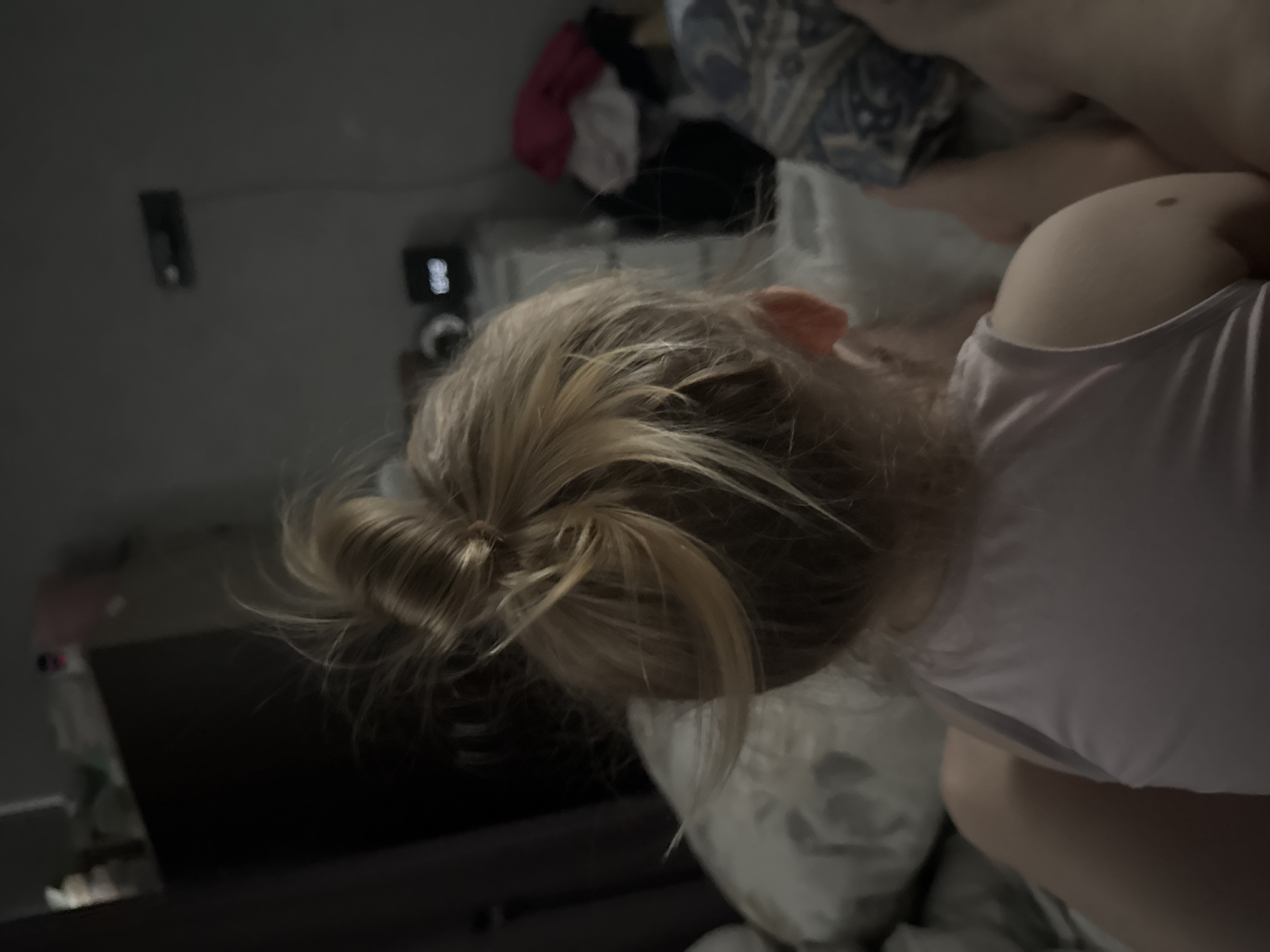 This media file shows a person lying down, with their head turned away from the camera. The person has blonde hair pulled back in a messy bun. They are wearing a light-colored t-shirt. The background appears to be a bedroom, with a bed and some clothing visible. There is also a dark surface that could be a television screen or a piece of furniture. The lighting is dim, suggesting it might be nighttime or a dimly lit room. No specific activity or event is clearly depicted, but the scene suggests a relaxed or resting state. There are no visible text elements in the image. The location context of Mikhaylovsk, Russia, provides no specific visual cues within the image itself.