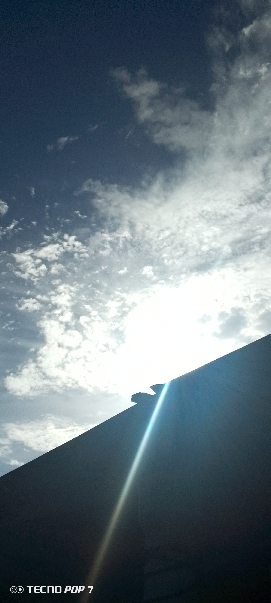 The image, taken in Altagracia, Venezuela, captures a view of the sky filled with scattered clouds, partially obscured by what appears to be the edge of a dark-colored building or structure in the foreground. The sun shines brightly from behind the clouds, creating a significant glare and a distinct ray of light that stretches diagonally across the structure. The sky transitions from a darker blue at the top to a brighter, cloudier area closer to the horizon. The bottom of the image is dominated by the dark silhouette of the building or structure. "TECNO POP 7" is visible at the bottom left corner of the image. There are no people or discernible interactions visible in the frame.