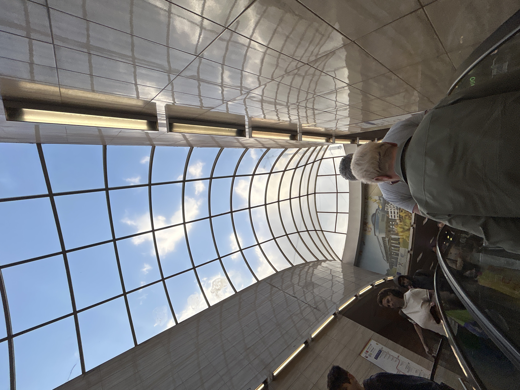 Captured from a tilted, low-angle perspective, the image shows people on an escalator under a transparent arched ceiling with a grid pattern, revealing a view of a blue sky with scattered clouds. The location is the Sofia Metro station in Sofia, Bulgaria, as indicated by the Cyrillic signs visible on the walls.
Fluorescent lights are installed above the ceiling, and a mural depicting a landscape scene is visible on the wall next to the escalator. Individuals on the escalator appear in various attire, some wearing jackets and shirts, with their faces partially visible. The escalator's handrail is black. The walls and ceiling are tiled.