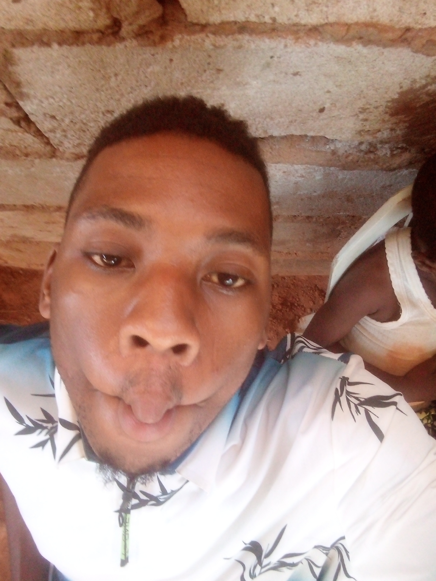 The image shows a man with short, dark hair and brown eyes, who is making a silly face with his tongue sticking out. He has a small beard and is wearing a light-colored shirt with a black leaf pattern. In the background, a person wearing a white tank top is partially visible. The location appears to be under a structure made of light-colored stone bricks. The city and specific location are Unknown, Unknown.