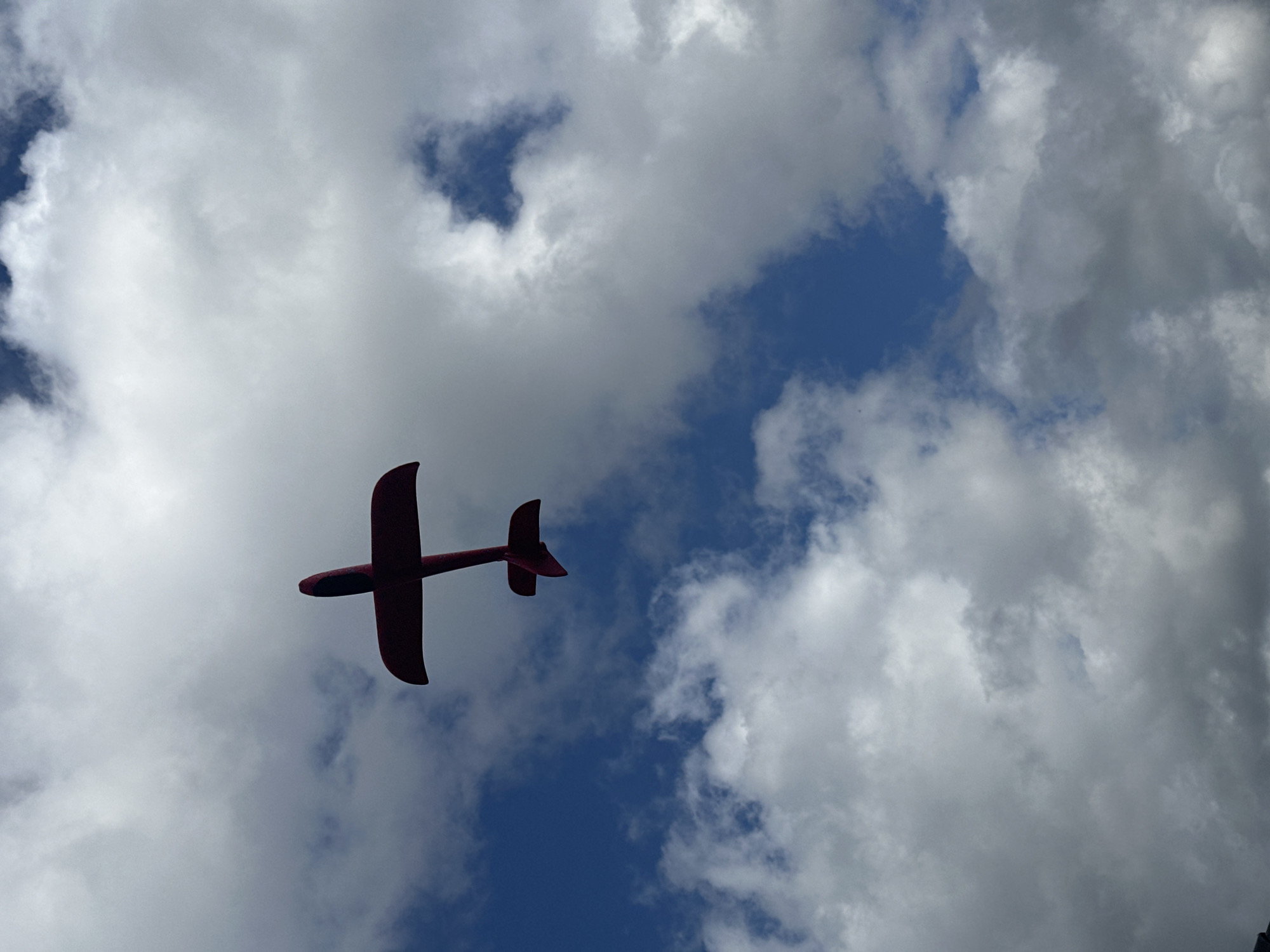 A low-angle shot from below captures a bright red, single-engine airplane soaring against a backdrop of blue sky and scattered white clouds. The aircraft is positioned in the upper center of the frame, viewed from its underside, with its wings spread wide and its tail feathers angled slightly upwards. The lighting suggests it is daytime, with sunlight illuminating the clouds. The image does not provide any specific details about the city of Wisła, Poland.