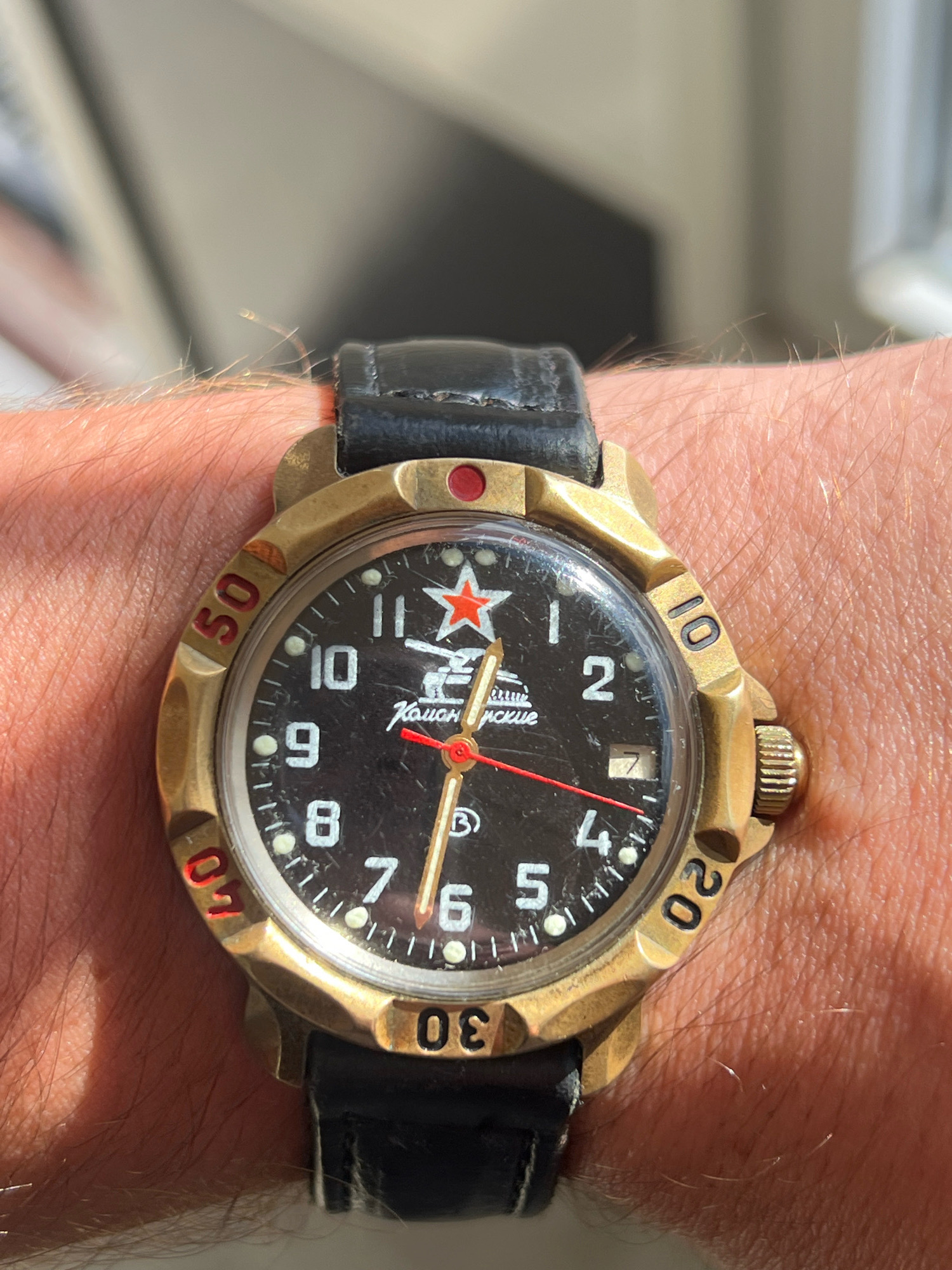 A close-up, eye-level shot shows a wrist wearing a watch. The watch has a black face with white Arabic numerals and a red star at the 12 o'clock position. The watch case is brass-colored, and the bezel has black markings. The watch band is black leather. The background is blurred, but appears to be an indoor setting with natural light. The location is Vilnius, Lithuania.