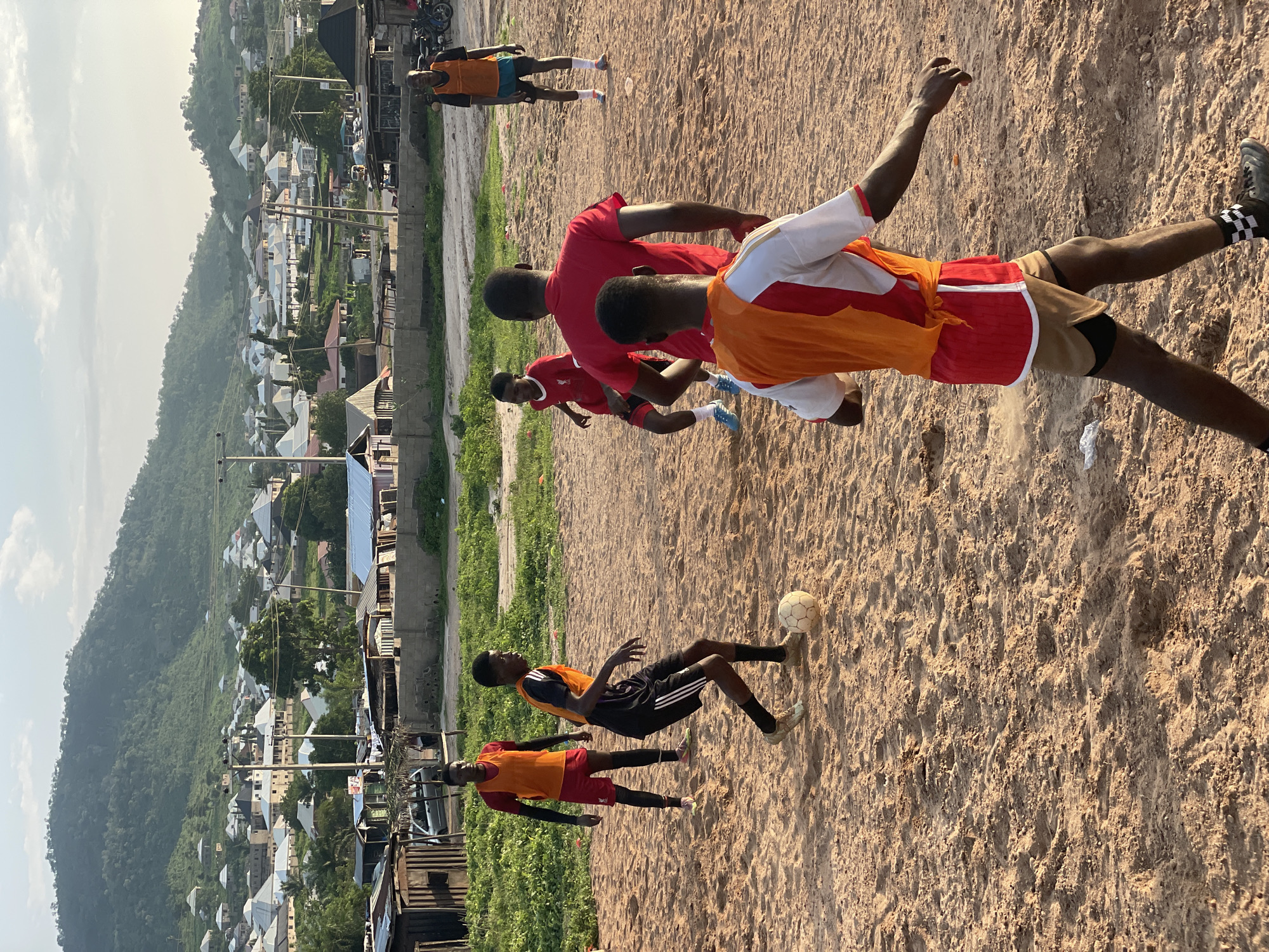 The image depicts a group of young men playing soccer on a sandy field in Lokoja, Nigeria. The sun is casting long shadows, suggesting it is either early morning or late afternoon. The sky is a pale blue with some scattered clouds, and the weather appears to be warm and clear.
The players are wearing casual athletic attire, including t-shirts and shorts. Some are wearing brightly colored bibs, indicating they are on opposing teams. One player in the foreground is in the process of kicking a soccer ball, with dust kicking up around his feet. Another player is running towards the ball, while others are positioned further down the field, anticipating the play.
In the background, a hillside settlement with numerous houses and buildings can be seen. The architecture suggests a developing area, with a mix of modern and traditional structures. Power lines crisscross the landscape, typical of many urban and semi-urban areas. The overall scene conveys a sense of community and recreation, with the young men engrossed in their game. There is no visible text in the image.