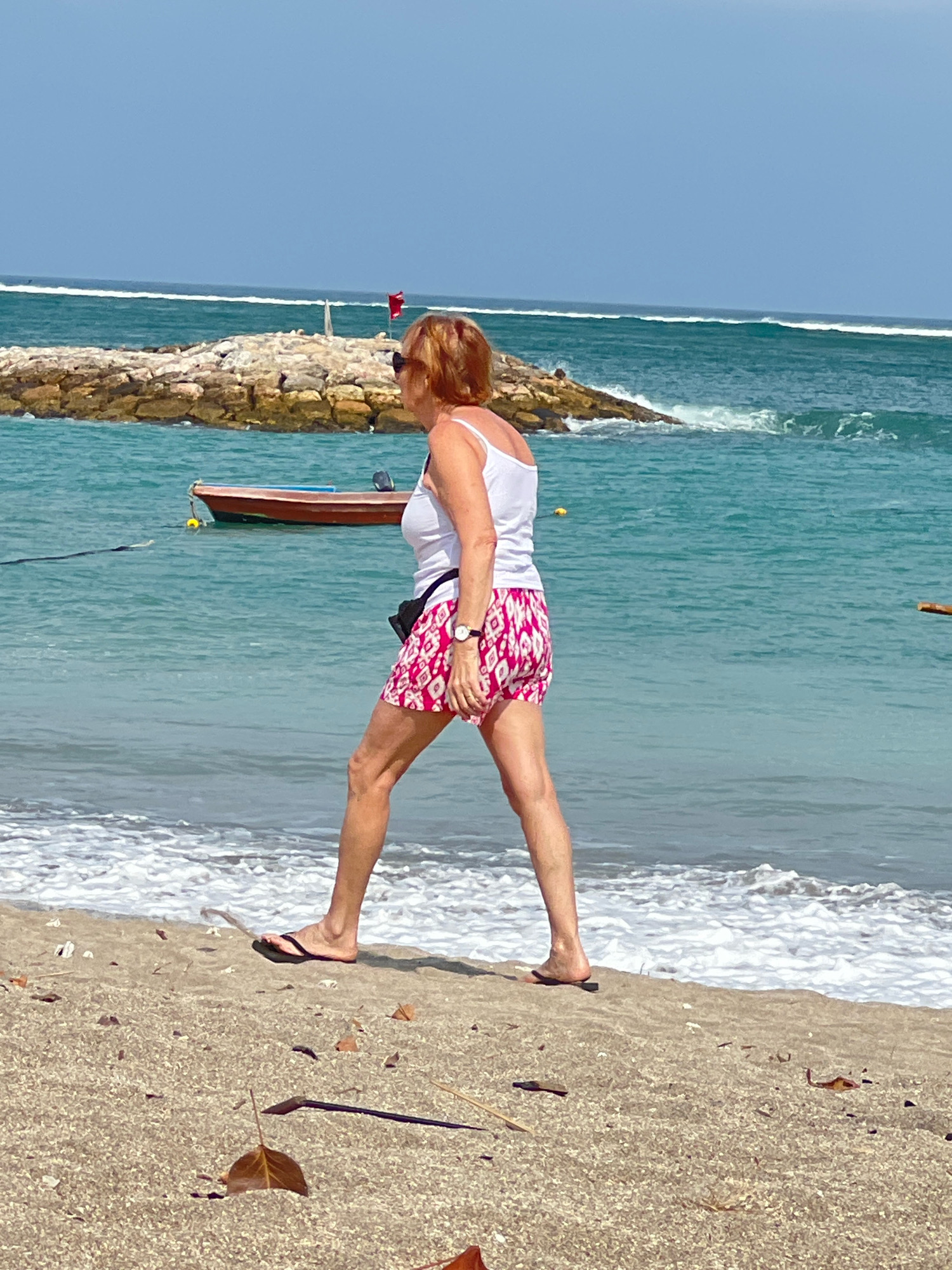 A woman with auburn hair, wearing sunglasses, a white tank top, and pink patterned shorts, walks along the sandy beach in Kuta, Indonesia. The ocean is a vibrant turquoise with white waves crashing near the shore. In the background, a stone jetty with a red flag and a small wooden boat moored to it are visible. The sky is clear blue. The sand is littered with dried leaves and debris.