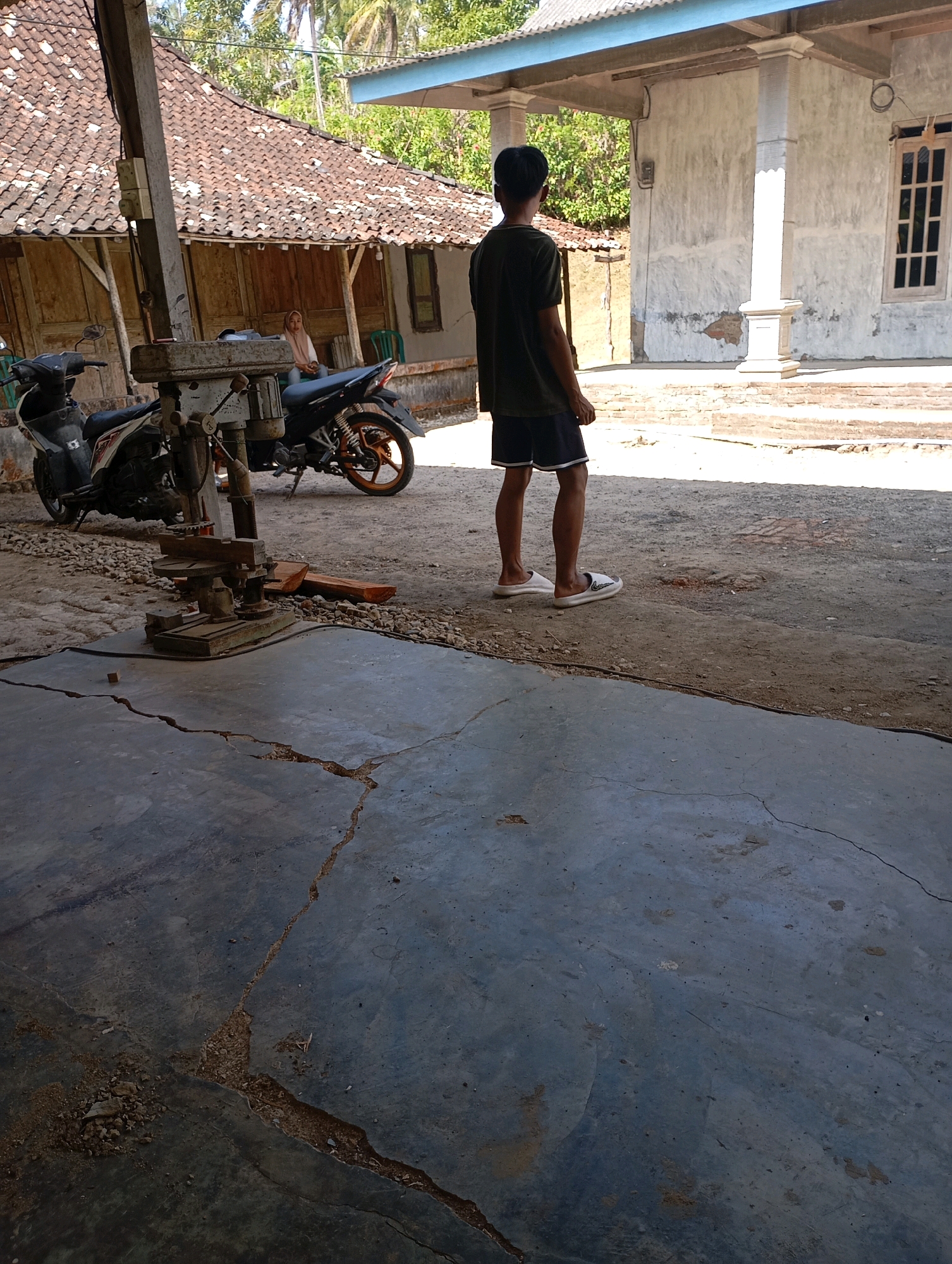A young man stands with his back to the camera, facing a dilapidated building with a corrugated metal roof and weathered wooden walls. He is wearing a dark green t-shirt and black shorts with white trim, and white slide sandals. Two motorcycles are parked to his left. The ground is a mix of cracked concrete and dirt. In the foreground, a rusty drill press sits on the concrete. In the background, a portion of a white building with a light blue overhang is visible. The scene appears to be an outdoor workshop or yard in a rural or semi-rural area of Ketepung, Indonesia. The light suggests it is daytime.