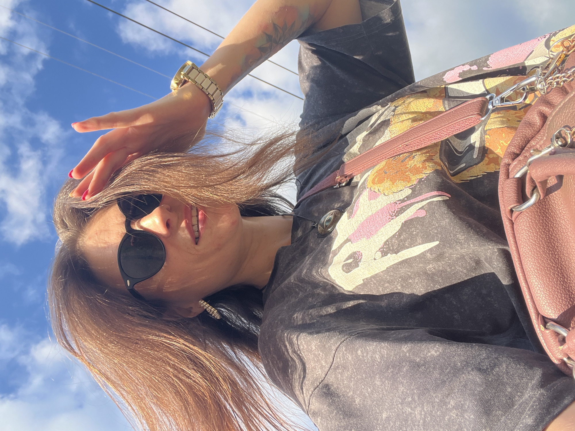A woman stands outdoors on a sunny day, with a blue sky and scattered clouds overhead. She is looking towards the camera and has her right hand raised, tucking her hair behind her ear. She is wearing sunglasses, a dark t-shirt with a graphic print, a pendant necklace, earrings, and a watch on her left wrist. A pink crossbody bag is visible in the foreground. The image appears to have been taken in Krasnoe-na-Volge, Russia.