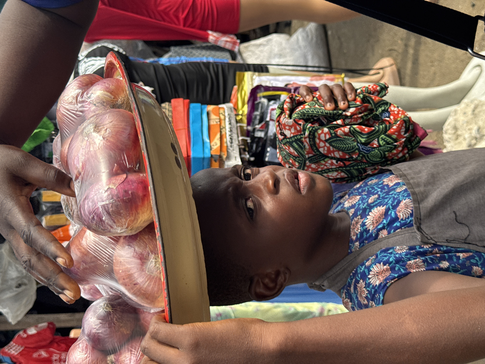 The image captures a candid, rotated view of a market scene, likely in Ado, Nigeria. A dark-skinned child, possibly a young girl, is prominent in the foreground, lying on her side and looking upwards with a neutral, contemplative expression. She wears a blue top adorned with white and orange floral patterns, layered under a plain grey garment. Her right arm supports a light-colored, elongated tray or board, while her left hand rests on a colorful, patterned fabric with motifs in red, green, white, and black.
On the tray, several red onions are neatly arranged, each individually wrapped in clear plastic. An adult's dark-skinned hands are visible on the far left, also supporting the tray and interacting with the packaged onions. The background reveals stacks of various market goods, including colorful fabrics or packaged items in shades of red, blue, orange, yellow, and purple, indicating a vibrant retail environment. A dark structural element is partially visible in the upper right. The diffused, soft lighting suggests daylight, possibly within a covered market area. No discernible text is visible within the frame. The scene portrays a moment of everyday life and activity in a local marketplace.