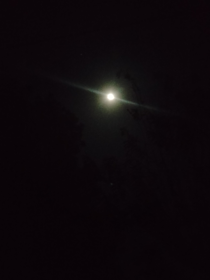 A dimly lit night sky displays a bright moon with a horizontal lens flare. Wisps of clouds partially obscure the moon's glow. Two smaller, faint points of light appear; one in the upper left quadrant, and another in the lower central area. The remainder of the image is predominantly dark, emphasizing the stark contrast between the light sources and the surrounding night.