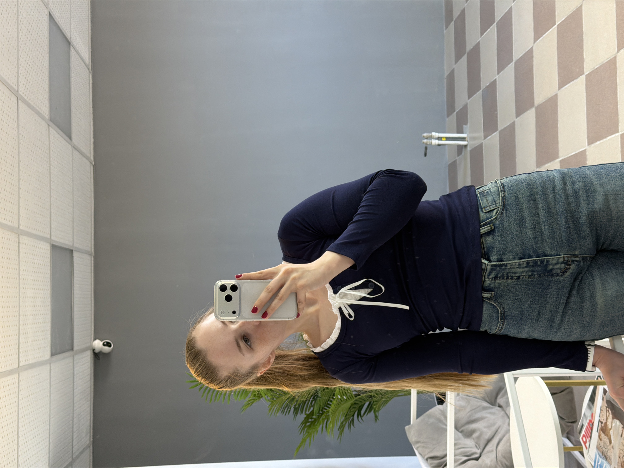 A woman with long, light brown hair is taking a selfie in a room with a neutral color palette. She is holding a silver smartphone in her right hand. The phone is positioned in front of her face, obscuring part of her expression. She is wearing a long-sleeved, navy blue shirt with a white, frilly neckline tied with ribbons. She is also wearing blue jeans. Behind her, there is a gray wall and a patterned, light brown and cream tile wall. A surveillance camera is mounted on the wall. A green, potted plant is visible near the bottom of the frame, and part of a white chair can be seen as well. The location is in Berezniki, Russia, inferred from the presence of specific details such as the tile pattern, wall color, and surveillance camera placement that are commonly seen in buildings of that region.