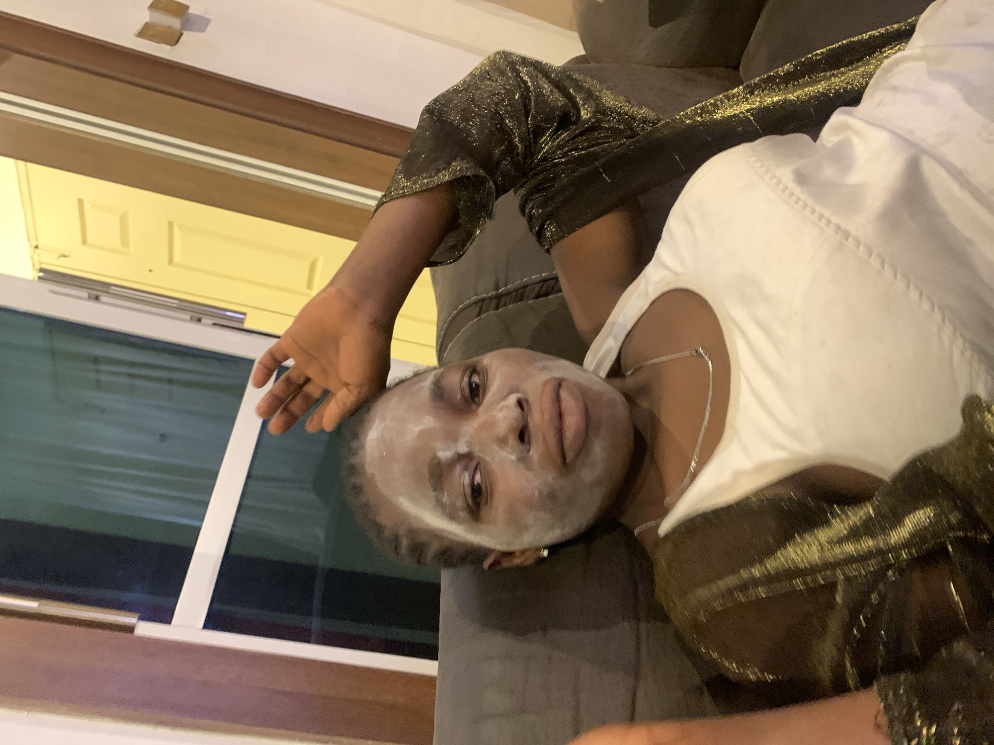 A person lies on a gray sofa. Their face is covered with a white substance. The person wears a white top and a black and gold top. A window is on the left side of the frame, and a door is near the top of the frame. The location is Jos, Nigeria.