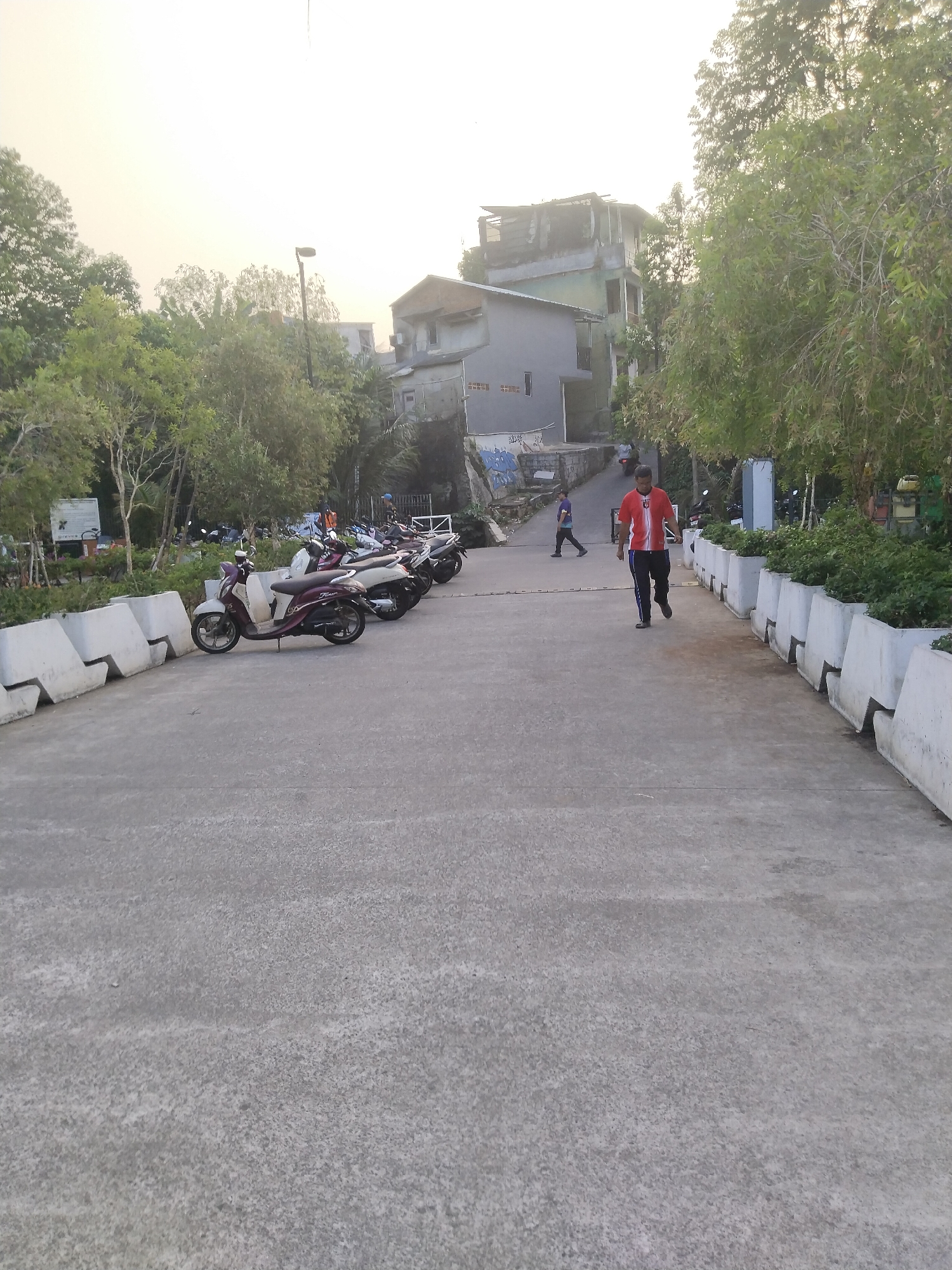 In an urban outdoor setting, a person in a red and white striped shirt and dark pants walks away from the camera on a concrete path. Another person, wearing a blue shirt and dark pants, is walking towards the camera in the background. The scene features several parked scooters on the left side of the path, beyond a concrete barrier. Buildings and trees are visible in the background, with a bright, overcast sky.