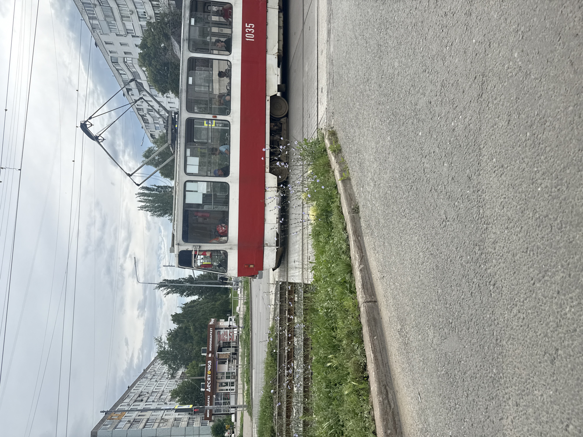 A tram, numbered 1035 and painted red and white, is moving right to left across the frame. It is on a tram track, with overgrown grass and a concrete curb to its left. In the background, a multi-story apartment building with many windows dominates the scene, with trees and other buildings visible further back. Overhead, power lines stretch across the cloudy sky. The tram has several passengers visible through its windows. The scene appears to be a residential area in the city of Samara, Russia, likely in a suburban or older part of the city given the architecture and condition of the tram tracks and curb.
