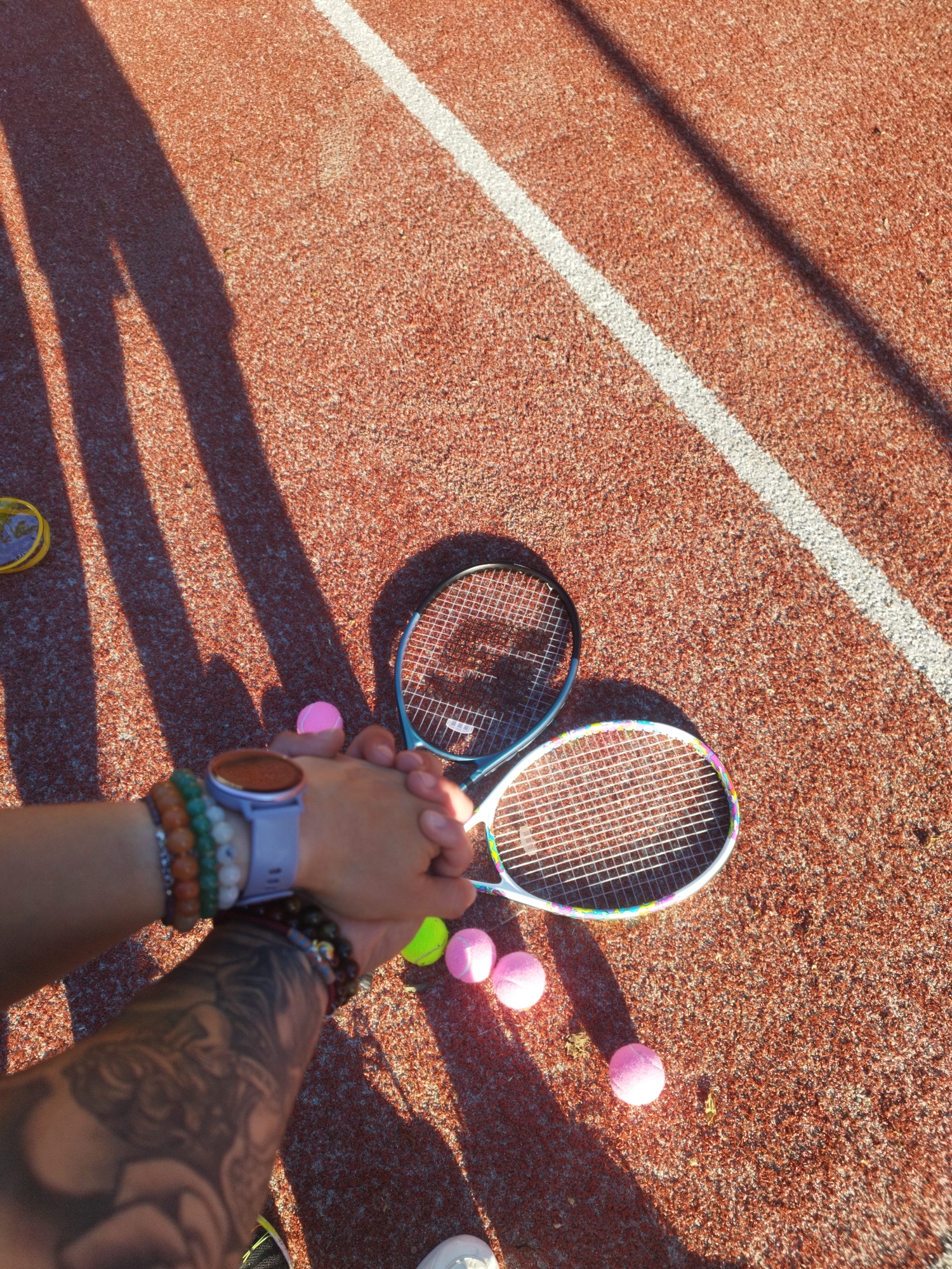 A high-angle shot shows two individuals grasping hands over two tennis rackets and scattered tennis balls on a reddish-brown surface with a white line. The surface is likely a tennis court or similar athletic surface. One arm exhibits a visible tattoo and a wrist adorned with beaded bracelets and a smartwatch with an orange face. The other wrist also has several beaded bracelets. The tennis rackets are different colors, one with a blue frame and the other white with a colorful rim. Pink tennis balls are spread around. The location is Lesidren, Bulgaria. The lighting indicates a sunny day, casting shadows.