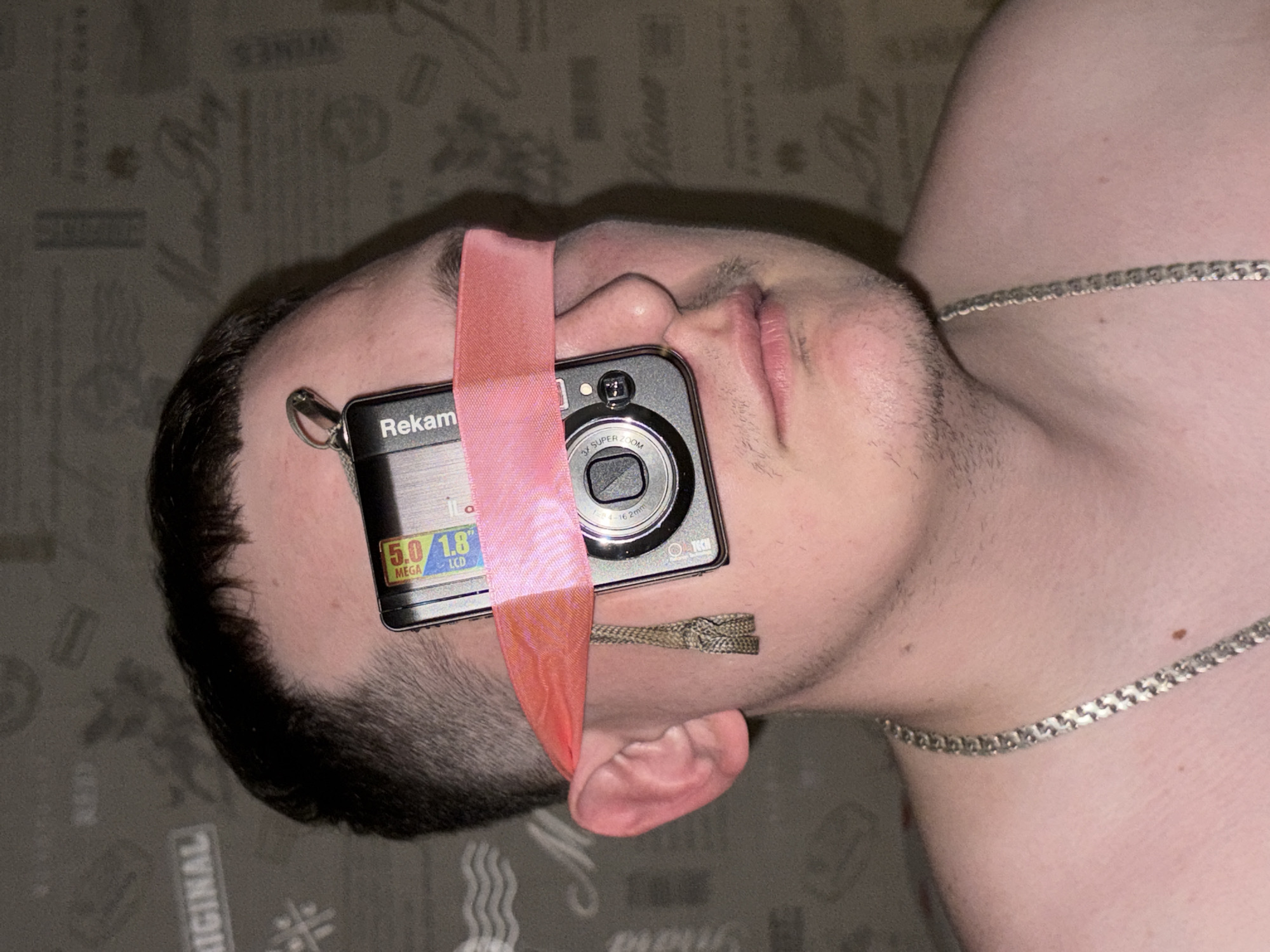 A man with short, dark hair is lying on a surface with a patterned, dark-colored covering. A silver chain necklace rests on his upper chest. A small, black Rekam digital camera is positioned over his right eye, obscuring it, and is held in place by a wide, light pink ribbon that extends from his forehead down to his ear. Part of a thin, braided cord is also visible near his ear. The location is not possible to determine.