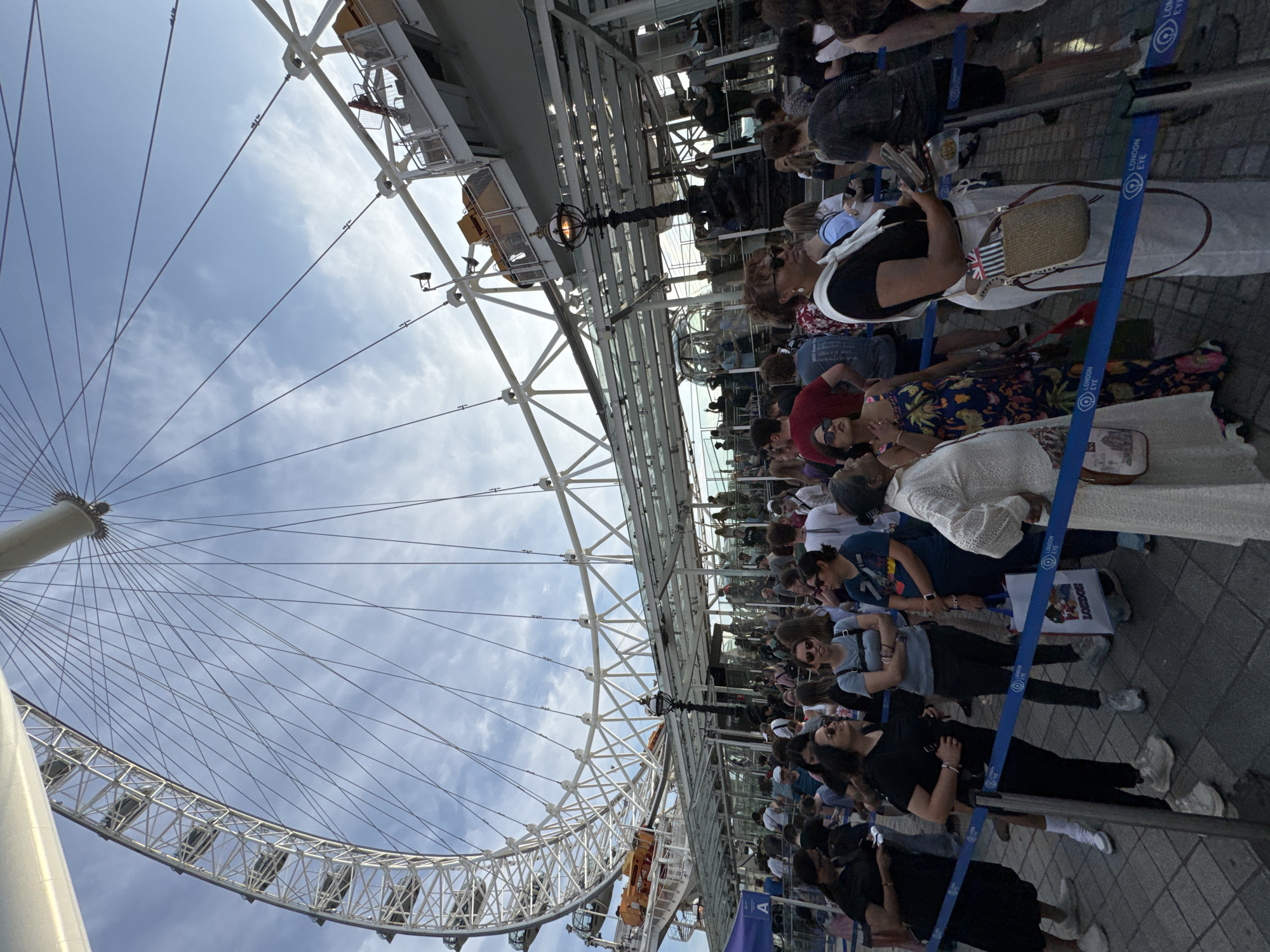 The image depicts a crowd of people queuing in front of the London Eye, a prominent landmark in London, United Kingdom. The large Ferris wheel, a signature structure of the attraction, dominates the upper portion of the frame. The queue is organized with blue stanchions and ropes, guiding visitors towards the entrance of the London Eye. People of various ages and ethnicities are visible, dressed casually for an outdoor excursion. Some are looking towards the attraction, while others are engaged in conversation or looking at their phones. The ground is paved with square tiles. The sky is partly cloudy, with a mix of blue sky and white clouds. The overall scene suggests a busy tourist attraction, likely during a popular time for visitors.