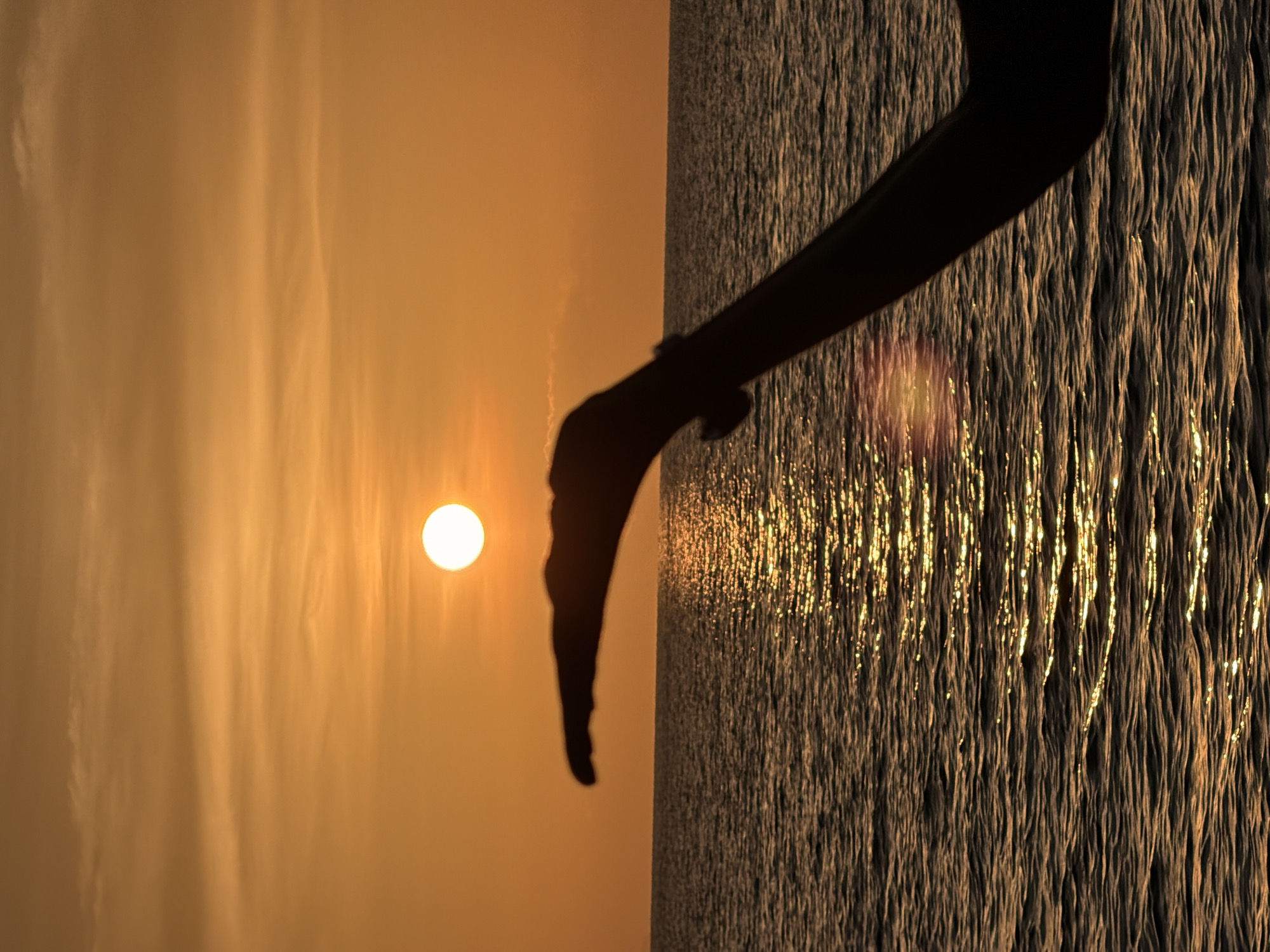 The image depicts a silhouetted arm and hand extended towards the ocean at sunset. The sun, a bright orb, is positioned in the upper center of the frame, casting a warm golden glow across the sky and reflecting on the rippling water. The hand is cupped, as if to catch the sun, creating an illusion that the sun is being held. The background is a gradient of orange and yellow hues, characteristic of a tropical sunset. The sea is calm with gentle waves, and the light creates a shimmering path across the water's surface. The scene is likely captured at a beach or a vantage point overlooking the sea in Ko Samui District, Thailand, a popular tourist destination known for its beautiful sunsets.