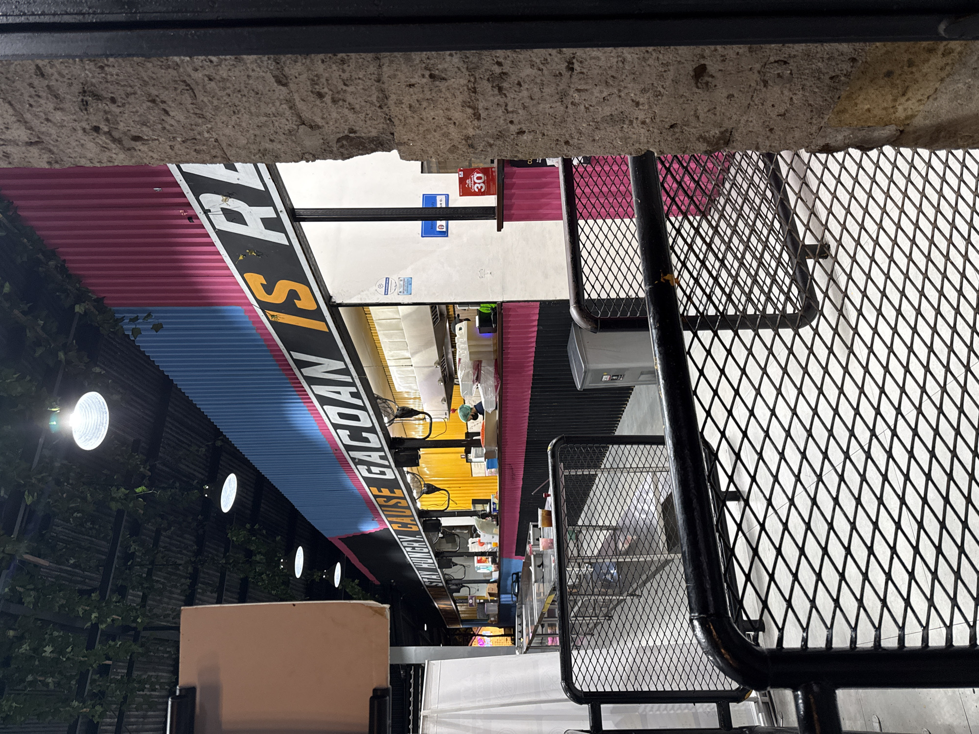 The image, taken in Kuta, Indonesia, captures an indoor commercial space, likely a food court or market street, illuminated by artificial lighting, suggesting evening or nighttime.
In the immediate foreground, a rough, grey concrete ceiling or beam runs across the top of the frame. Below it, a black metal structure, possibly part of a table, chair, or partition, features a diamond-patterned mesh.
The scene beyond the foreground elements depicts a vibrant row of shopfronts or stalls. To the left, a wall covered in green foliage is visible, with several bright, circular lights illuminating the area. The stalls feature corrugated metal panels in various colors, including prominent pink and blue sections. A long, black signboard with white and yellow lettering stretches across the midground. The visible text on this sign reads: "STAY HUNGRY CAUSE GACOAN IS RE..." with the final word partially cut off. Further into the background, other stalls are visible with yellow and pink elements, and another red sign displays the number "30". No people are clearly visible as primary subjects. The general setup suggests an active commercial environment, possibly offering food, typical of a busy market or food court setting.