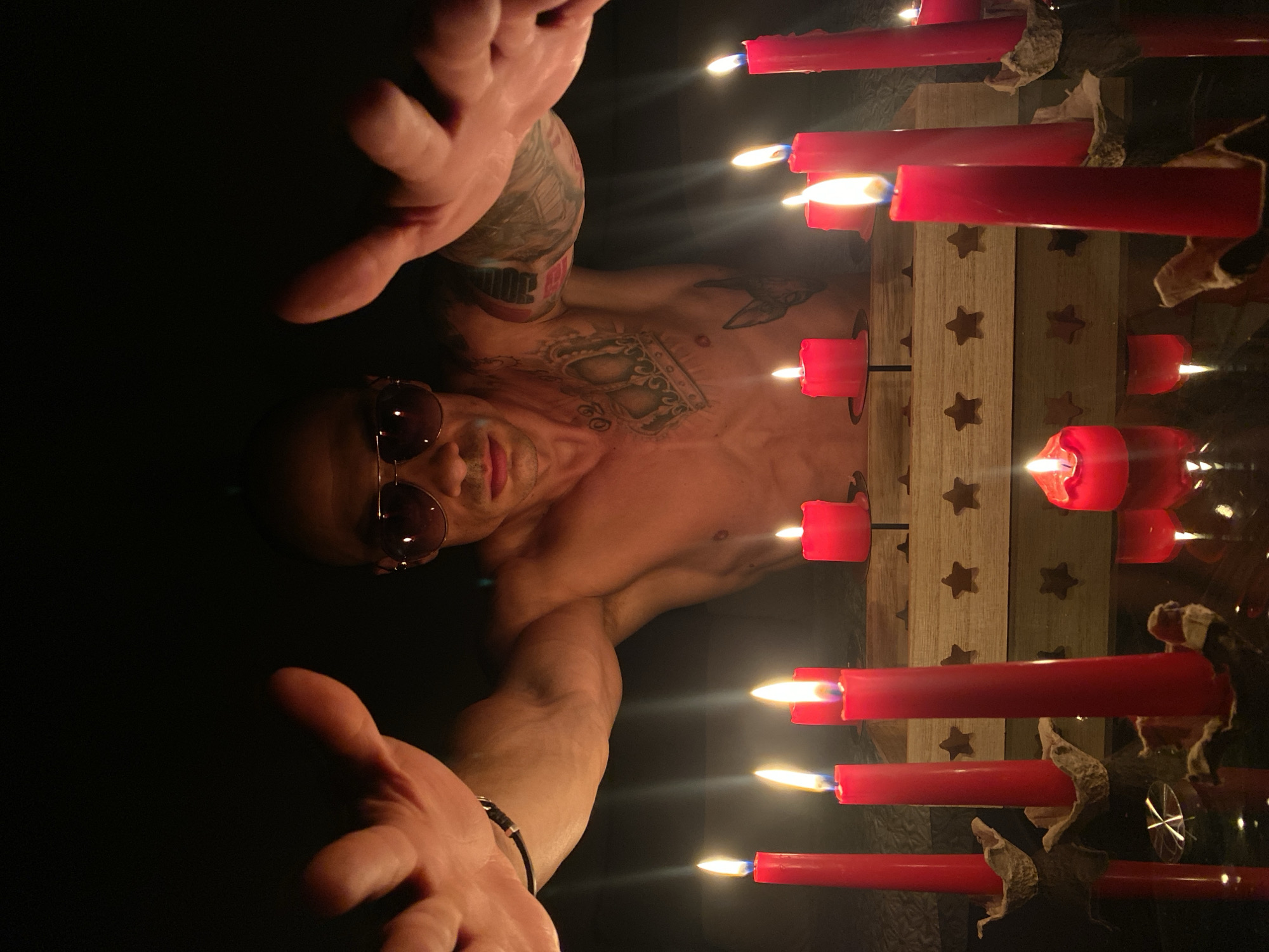 The image shows a man with tattoos on his chest and arm, wearing sunglasses. He is shirtless and his hands are outstretched towards the camera. In front of him are several red candles, some of which are lit. The candles are placed on a reflective surface, possibly a table, and a wooden holder with star cutouts is visible in the background. The overall setting is dimly lit, with the flames of the candles being the primary light source. The man appears to be in a room, but the background is mostly dark and indistinct. There is no information in the image that places the scene in the city of Pleven, Bulgaria.