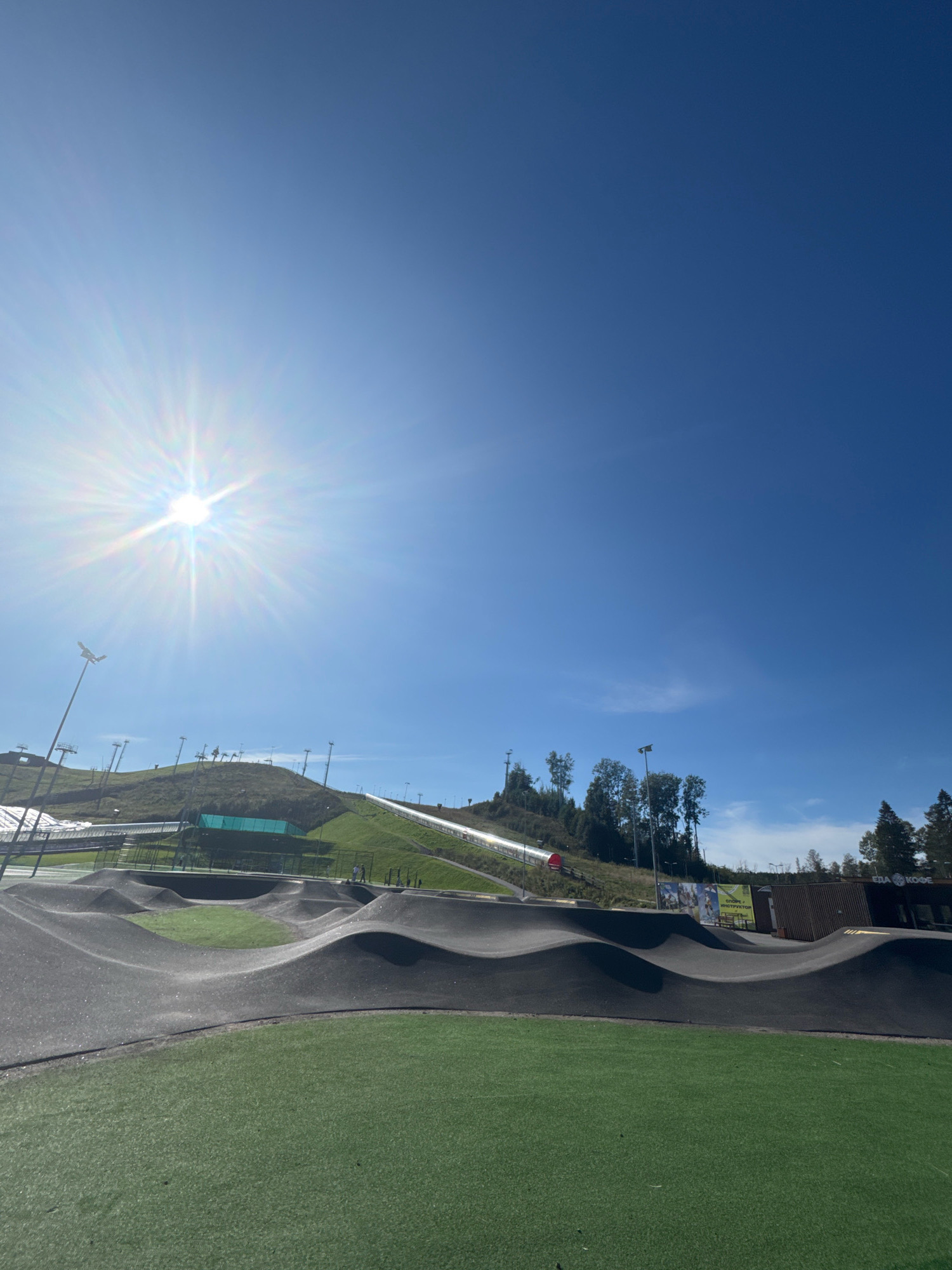 In Okhtinskiy Park, Russia, a sunny day illuminates a vibrant outdoor recreational area. The foreground features a professionally designed pump track, characterized by its smooth, undulating asphalt surface with integrated green turf zones. Beyond the pump track, the terrain slopes upwards, revealing a ski jump with a metallic track and surrounding grassy slopes dotted with numerous floodlights. Trees sparsely populate the hillside. In the mid-ground, two individuals are visible near the base of the ski jump, while a small wooden building with signage reading "Еда и Кофе" (Food and Coffee) stands to the right. The sky is a clear, bright blue.