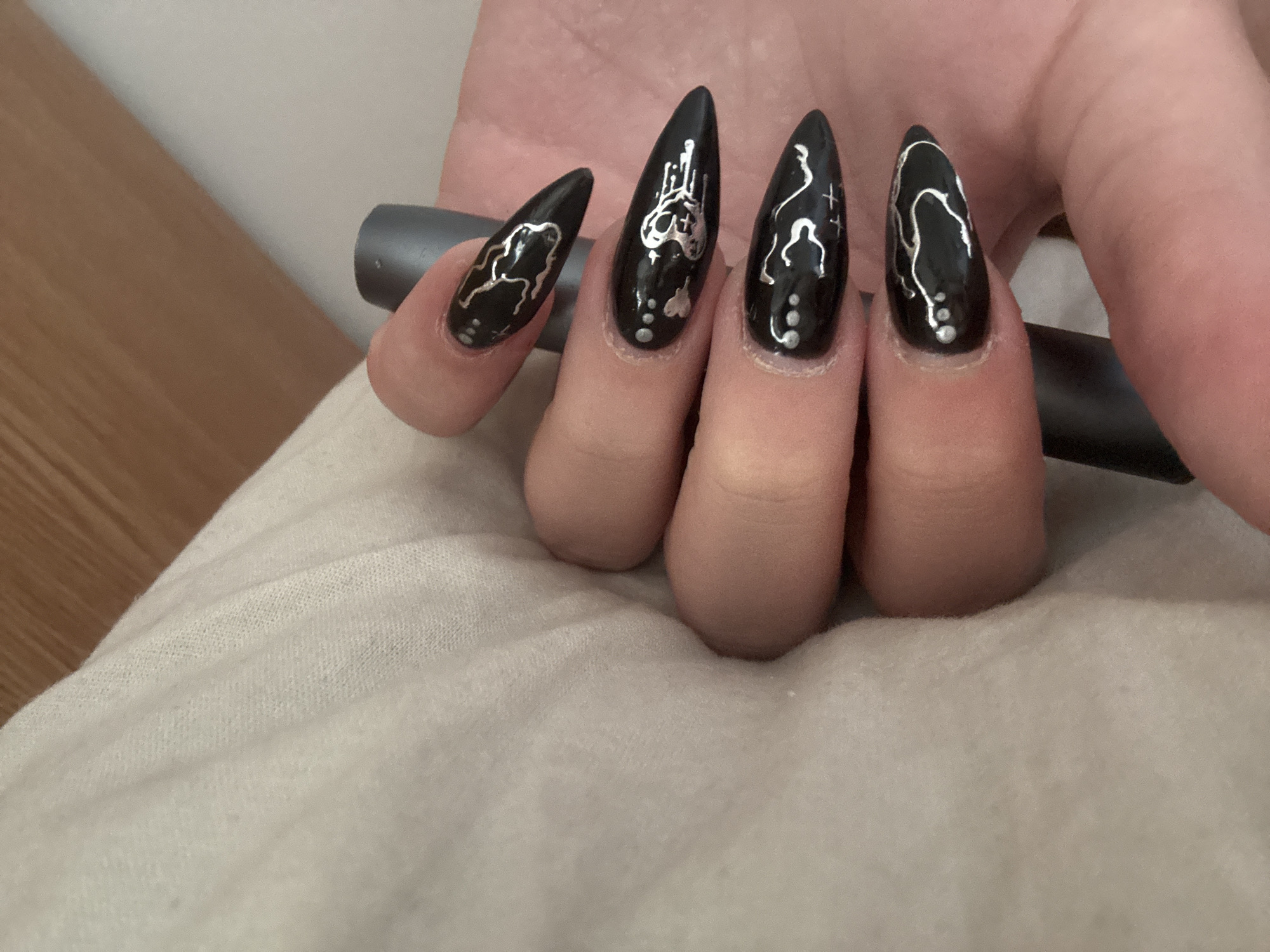 A close-up image shows a person's hand holding a grey cylindrical object. The fingernails are long and pointed, painted black with silver designs. The designs are abstract, with dots near the base of each nail. The hand rests on a light-colored, textured fabric, possibly a blanket or sheet. The location, Troyan, Bulgaria, is not evident in the image.