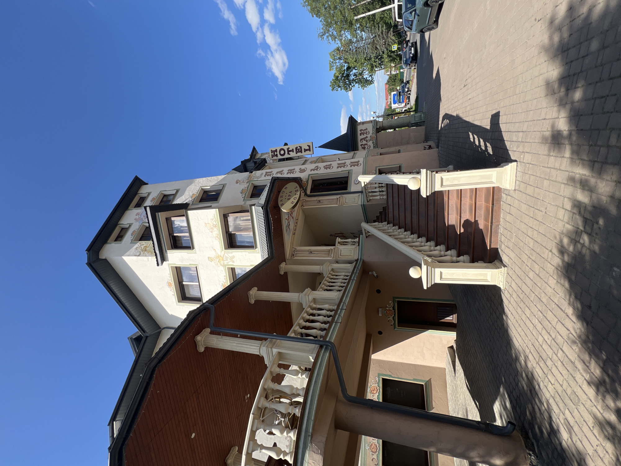 An exterior, low-angle, wide shot captures the facade of a hotel in Yaremche, Ukraine, under a clear blue sky with scattered clouds. The hotel is a multi-story building with a light-colored exterior adorned with decorative painted murals and ornate trim. A wooden-covered balcony with a white balustrade extends from the second story. A set of brown-treaded stairs with white balustrades leads up to the hotel entrance. A vertical "HOTEL" sign is mounted on the side of the building, and another smaller sign reads "BARON SPA HOTEL". The hotel is situated on a paved street with parked cars and trees visible in the background. The pavement in front of the hotel is laid with grey bricks, and shadows from the building and trees are cast across it.