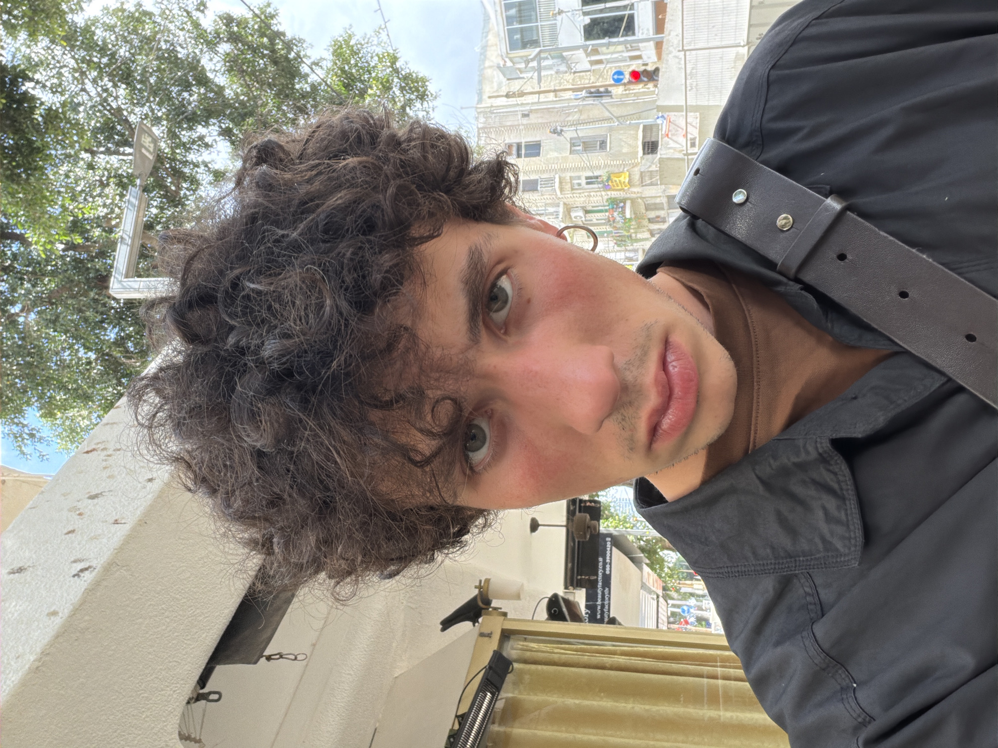 A selfie taken in Tel-Aviv, Israel. The subject is a young adult with curly brown hair, fair skin, and green eyes. They are wearing a dark-colored shirt and a brown leather belt is visible. The background includes buildings and trees, suggesting an urban environment. A traffic light is visible on one of the buildings. The photo has been rotated and appears to have been taken from a low angle.