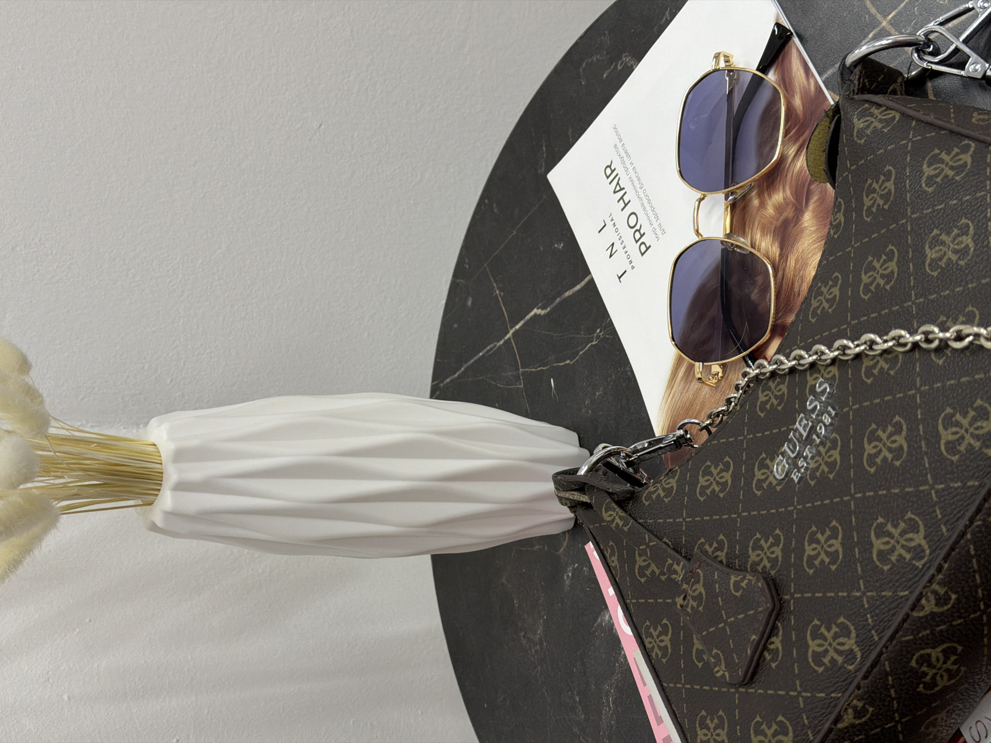A high-angle close-up shot shows a black marble-patterned table, angled slightly to the right. On the table are various items including a white vase with dried floral stems, a magazine advertising hair products with the words "TNL PRO HAIR PROFESSIONAL" prominently displayed, a pair of gold-framed sunglasses, and part of a brown patterned Guess brand purse with a silver chain strap. The background includes a section of white wall. The scene is well-lit, providing clear details of each item.