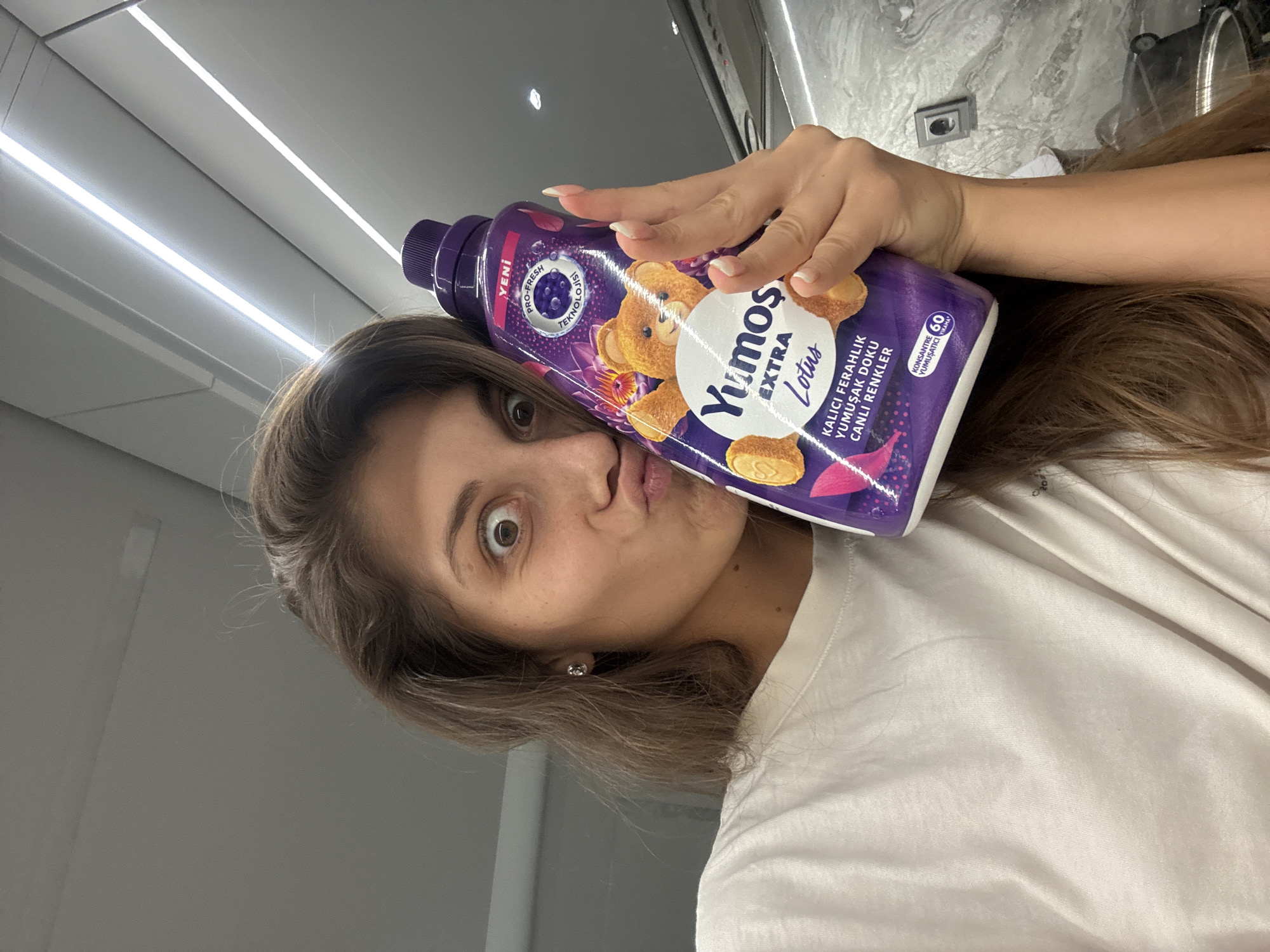 A woman with long brown hair is holding a purple bottle of Yumoş Extra Lotus fabric softener and making a kissing face. The bottle has a teddy bear on it and the text "KALICI FERAHLIK YUMUŞAK DOKU CANLI RENKLER." The woman is wearing a white t-shirt, and the background appears to be a kitchen with fluorescent lighting. It is impossible to pinpoint Burgas as the location with the information provided.