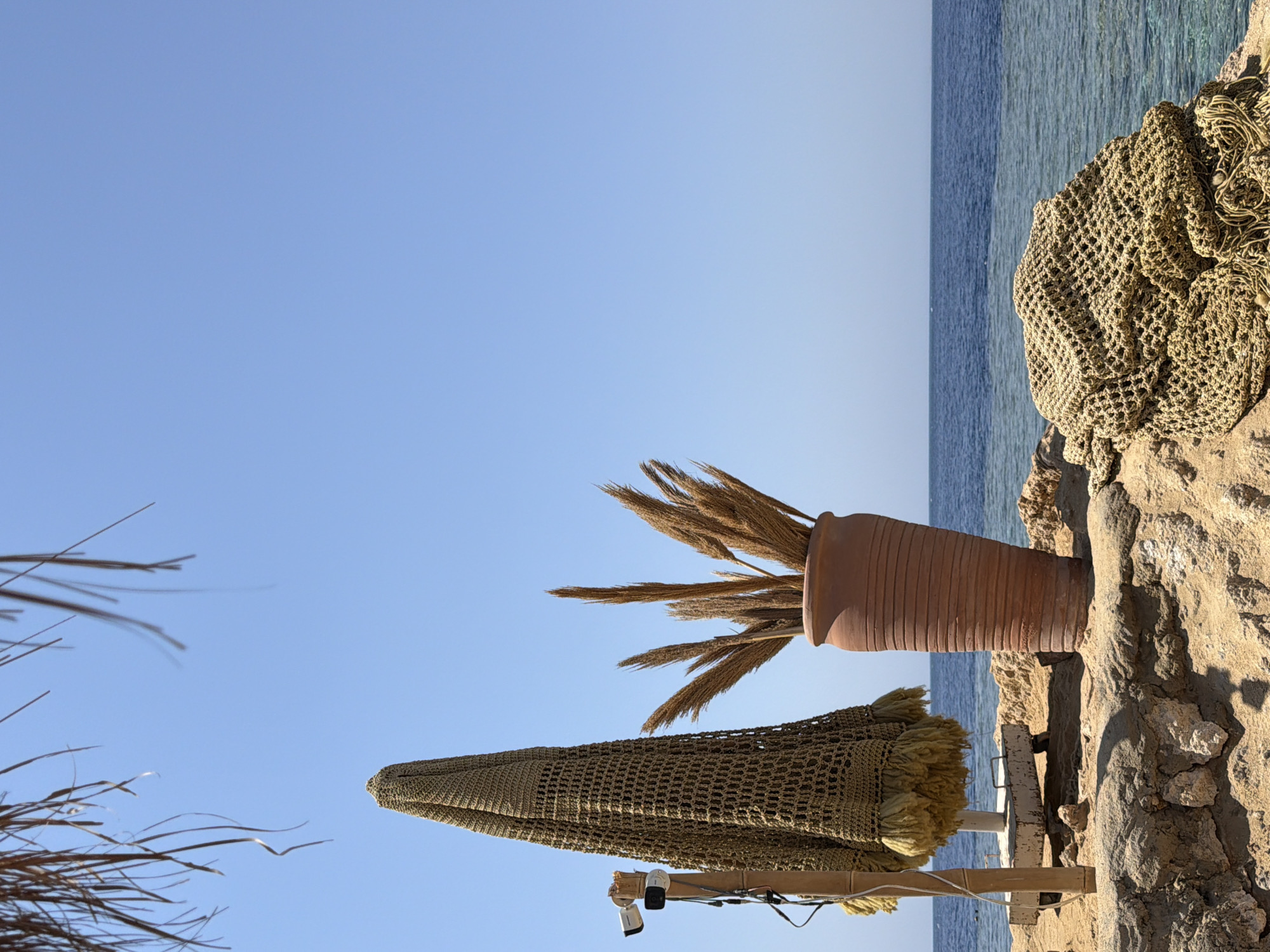 A still, medium shot captures a section of a beach on a sunny day with a clear blue sky and calm blue water. In the foreground, sand and rock formations are visible. To the left, a closed beach umbrella with a crocheted cover stands upright, supported by a wooden pole with a security camera attached. To the right of the umbrella, a large, terracotta vase holds a bouquet of dried reeds. In the mid-ground, a pile of netting rests on rocks. The background shows the expanse of the sea meeting the horizon under the bright sky. This scene is representative of a resort area in Sharm El Sheikh, Egypt.