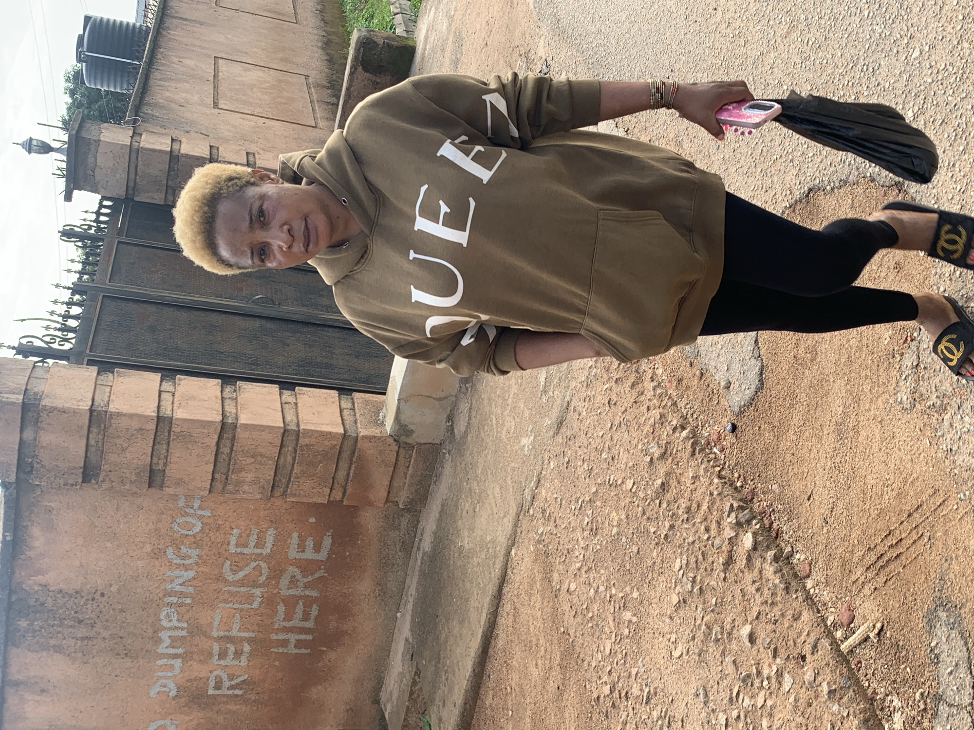 A woman with short blonde hair stands facing forward and slightly to the right, looking at the camera. She is wearing a khaki hoodie with the word "QUEE" printed in large white letters across the front. She is also wearing black leggings and black slides with gold logos. In her right hand, she holds a pink phone and a black plastic bag. The background consists of a textured, reddish-brown wall with white graffiti that reads "NO DUMPING OF REFUSE HERE." Behind the wall, a dark metal gate is visible. The ground in front of the wall is a mixture of dirt and small stones. The scene appears to be outdoors, possibly on a street in Jos, Nigeria, given the presence of the wall and the general environment.