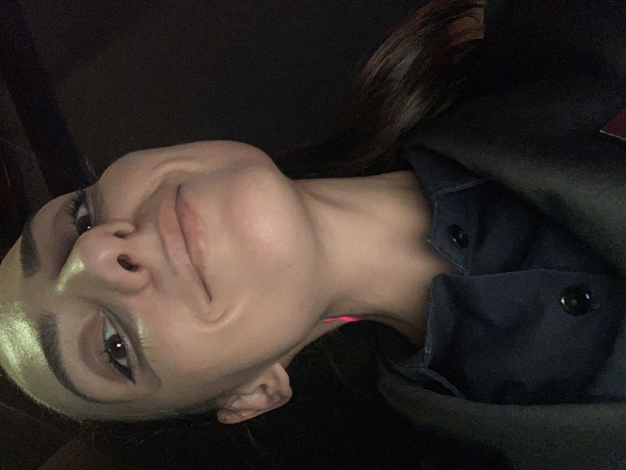 The image is a close-up, low-angle selfie of a young woman. She is looking up at the camera with a slight smile, her head tilted. Her makeup includes winged eyeliner and mascara. She is wearing a dark blue collared shirt with buttons and a dark jacket or coat. A thin red light is visible on her neck. The background is dark and appears to be indoors. No specific location in Burgas, Bulgaria is identifiable.