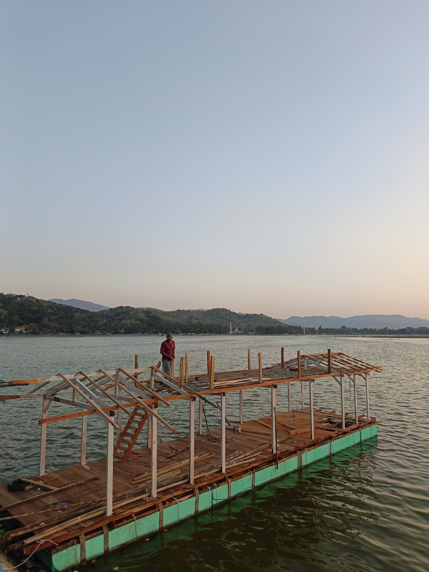 In Krakitan, Indonesia, a person wearing a red shirt and gray pants is standing on a wooden platform in a body of water. The platform appears to be under construction, with exposed wooden framing for a roof and a small ladder. The platform is supported by green flotation devices. In the background, a calm body of water stretches towards a shoreline lined with trees and hills. The sky above is a clear, pale blue, transitioning to a warmer hue near the horizon, suggesting either sunrise or sunset. The overall scene depicts a construction project in a serene, natural environment.