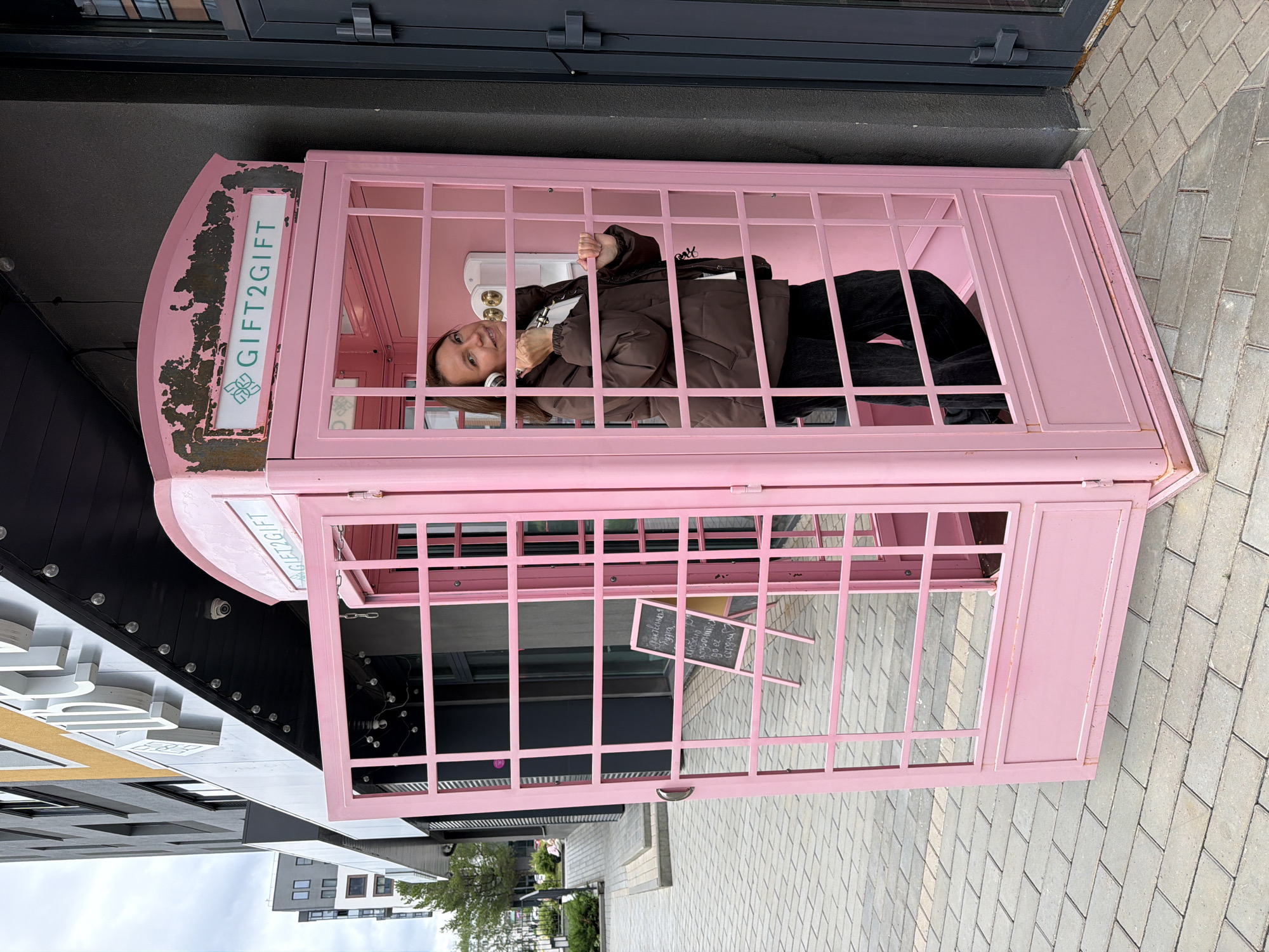 This image captures a wide outdoor shot of a woman interacting with a prominent pink phone booth on a paved sidewalk in Izhevsk, Russia. The woman, appearing to be middle-aged with dark brown hair, stands inside the pink phone booth. She is wearing a dark brown puffer jacket, a white shirt, and dark pants. Her left hand holds a white telephone receiver to her left ear, while her right hand rests on an interior horizontal bar. She looks directly at the camera with a slight smile.
The pink phone booth, reminiscent of a classic British design, features an open grid-patterned front door, which is swung open to the left. The top of the booth shows signs of wear, with chipped paint revealing rust-colored patches. A sign on the upper part of the booth displays "GIFT2GIFT" in teal letters with a corresponding logo. Inside the booth, a white rotary-style phone is mounted on the back wall.
The environment consists of a light gray rectangular paver sidewalk, which extends into the mid-ground. To the immediate right of the phone booth is a dark gray building wall with a dark door. To the left, a modern multi-story building with a facade primarily in gray and black, accented with yellow/ochre, stands behind the phone booth. White 3D lettering is visible on the building's exterior, and string lights are suspended under its overhang, where a small security camera is also present. In the background to the left, further urban buildings, a ramp, and some greenery, including a small tree and bushes, are visible under an overcast sky. Partially obscured behind the open phone booth door is a dark A-frame chalkboard sign with white Cyrillic writing.