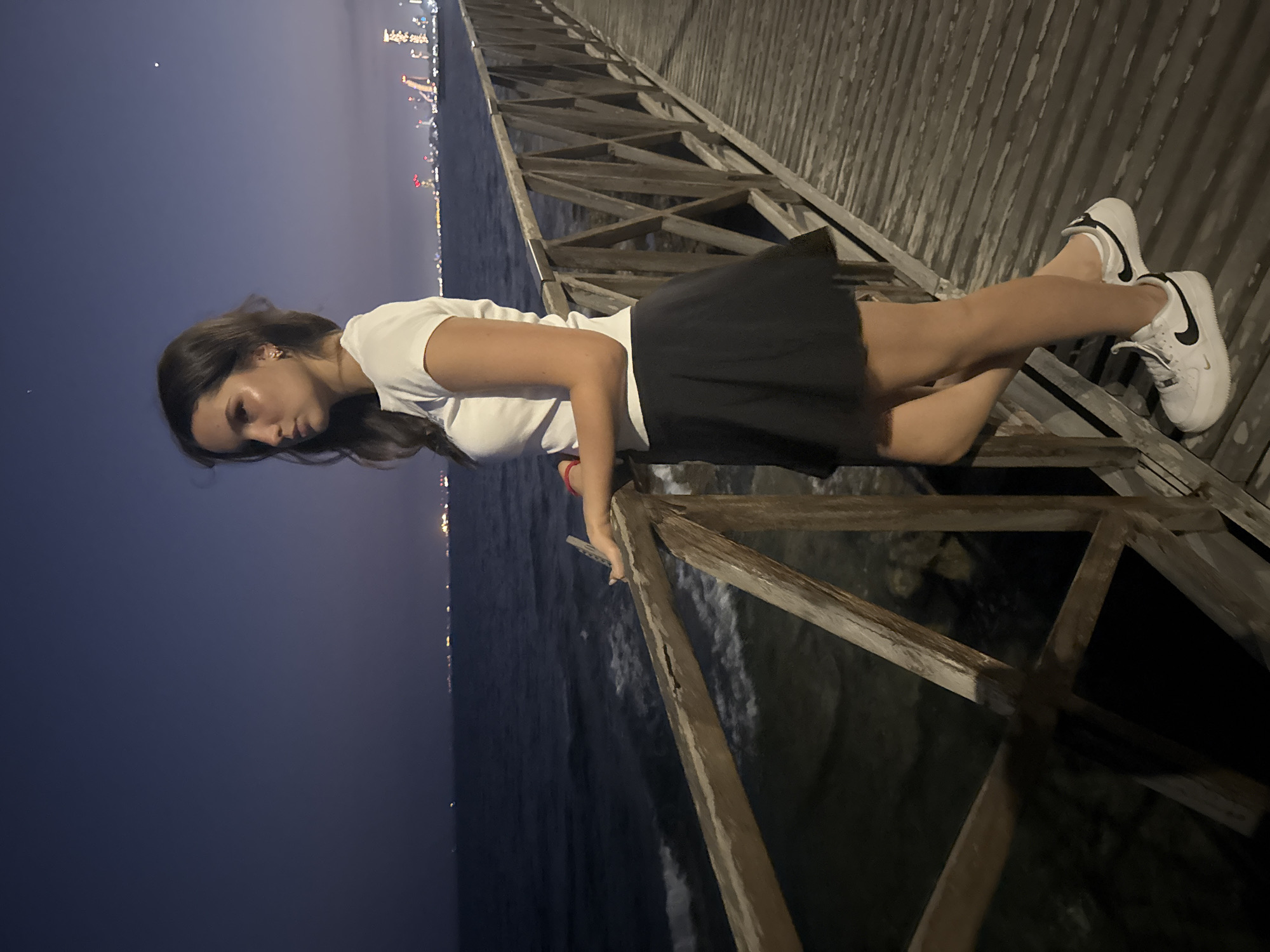 A young woman stands on a wooden pier at night. She is wearing a white t-shirt, a black skirt, and white sneakers. She is looking out at the ocean, with a city skyline visible in the distance. The water is dark, and there are small waves. The sky is a deep purple, with a few stars visible. The pier is made of weathered wood and has a simple wooden railing. The city lights are a mix of white and yellow. The location appears to be Agios Tychona Community, Cyprus.