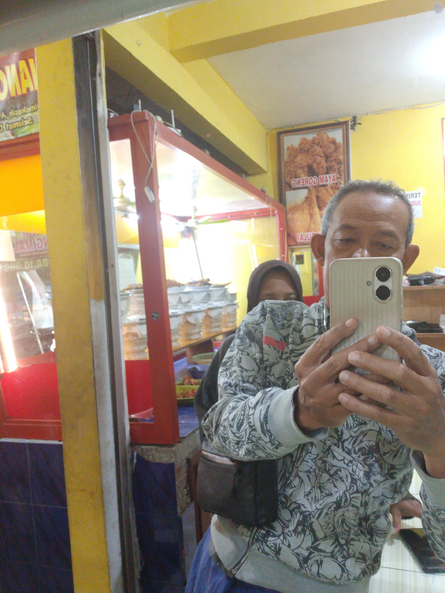 A man is taking a selfie in a room that appears to be a food stall or restaurant. The man is wearing a grey hooded sweatshirt with a black and white pattern and holding a silver smartphone in his right hand, angled to capture his reflection. Behind him, slightly out of focus, a woman wearing a brown hijab is visible.
The environment includes a bright yellow wall, a red-trimmed display case filled with food items, and a framed picture on the wall showing a pile of fried chicken with text that reads "AYAM GORENG MAYA" (fried chicken Maya). There is also signage visible on the left with partially obscured text. The floor has blue and white tiles. The lighting suggests an indoor setting, likely during the day or under artificial lights. The overall impression is a casual snapshot taken in a commercial food establishment.