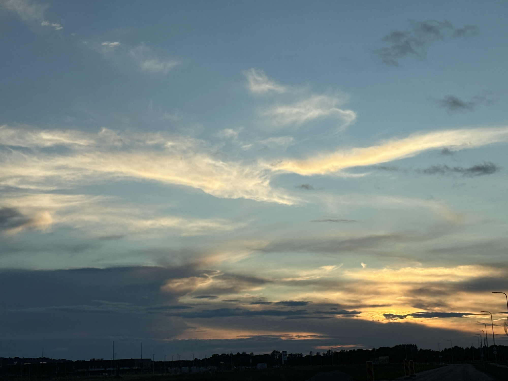 A sunset view over Tiskre, Estonia, captures a sky filled with wispy clouds tinged with orange and yellow hues, transitioning into darker blues and grays towards the horizon. The foreground is dominated by a silhouetted landscape featuring a dark line of trees and scattered buildings. Several lampposts are visible along what appears to be a road or pathway. The overall scene is serene, with the last light of day painting the sky.