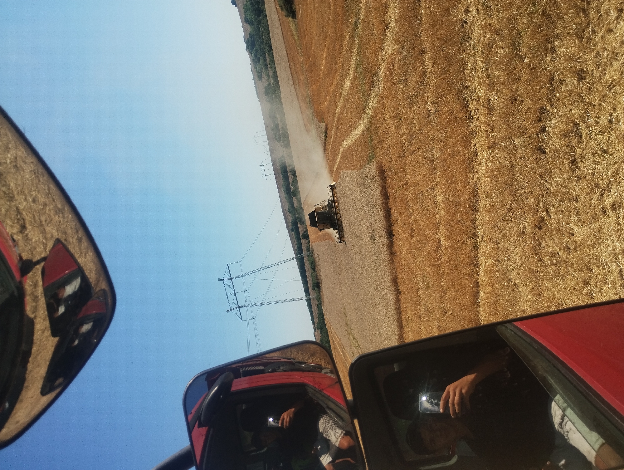 This image, taken in the city of Kubadin, Bulgaria, provides a glimpse of agricultural activity under a clear blue sky. The foreground features the reflection of a person taking a selfie in a red vehicle's side mirror. The person, wearing a dark t-shirt and light-colored shorts, is holding a smartphone. In the middle ground, a combine harvester is actively working in a golden field of harvested wheat, kicking up a cloud of dust as it moves. In the background, a set of large electrical pylons stands tall against the horizon, with rolling hills and scattered trees forming the distant landscape. The field itself is a patchwork of cut stubble and standing grain, indicating the ongoing harvest.