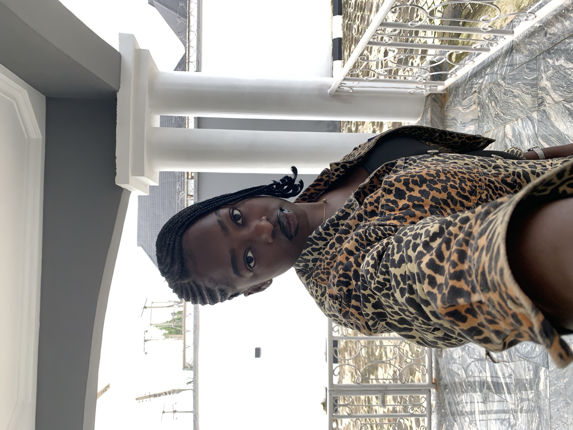 In the city of Gura Top, Nigeria, a young Black woman with braided hair and dark lipstick takes a selfie. She is wearing a leopard-print jacket over a black top. She is standing on a marble-tiled patio, which is bordered by a white metal railing and ornate columns. The background includes a white wall and structures that suggest a residential or commercial area.