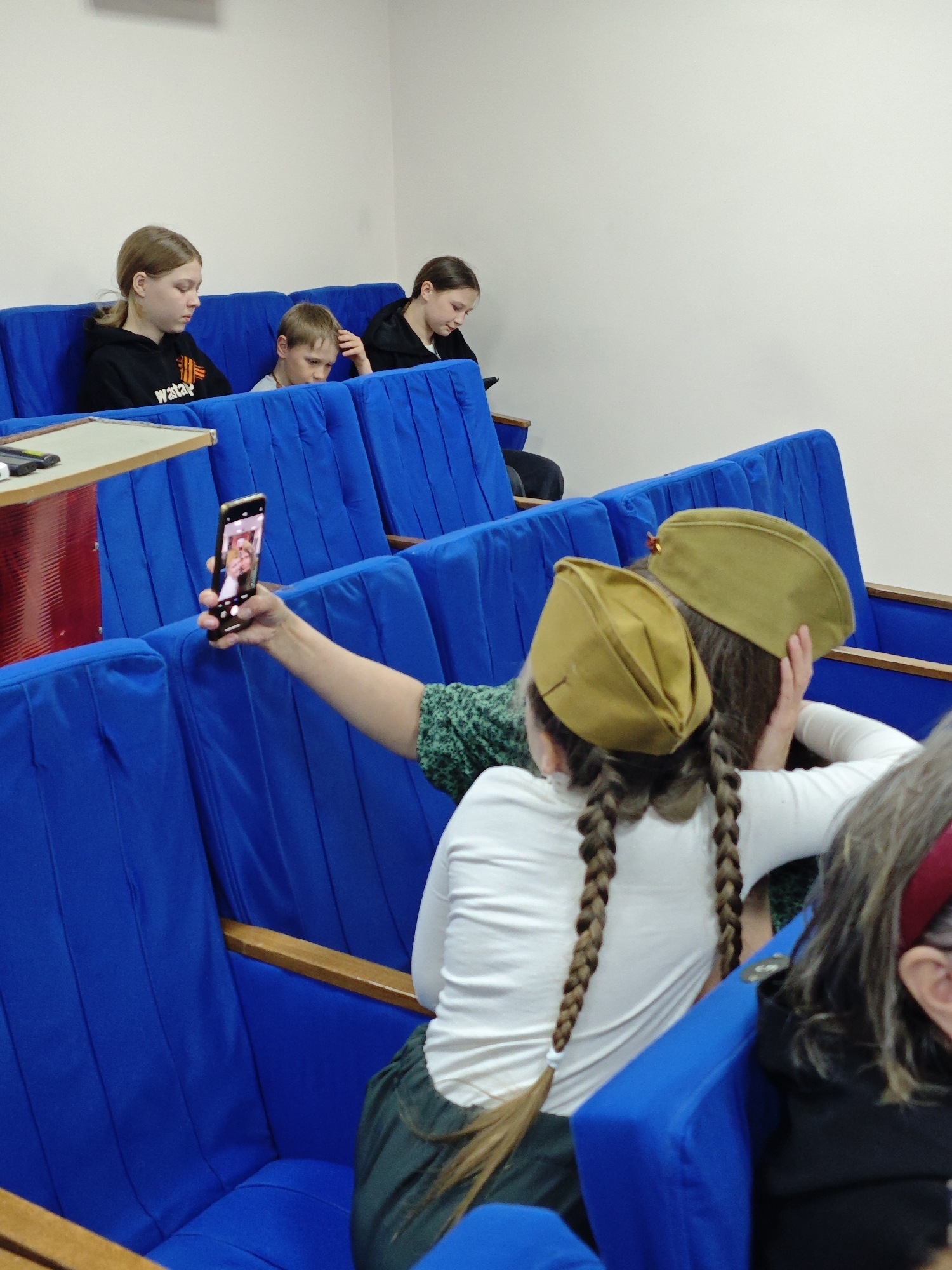 In Фоки, Russia, a group of people are seated in blue upholstered chairs, arranged in tiered rows suggestive of an auditorium or theater. A woman in the foreground, viewed from the back, extends her arm to take a selfie with a smartphone. She and another person next to her wear khaki-colored military-style caps. The woman has long braids. In the background, two girls and a boy are seated. One girl wears a black hoodie with a red and orange design. A table with items on it is to the left. The room has plain white walls.