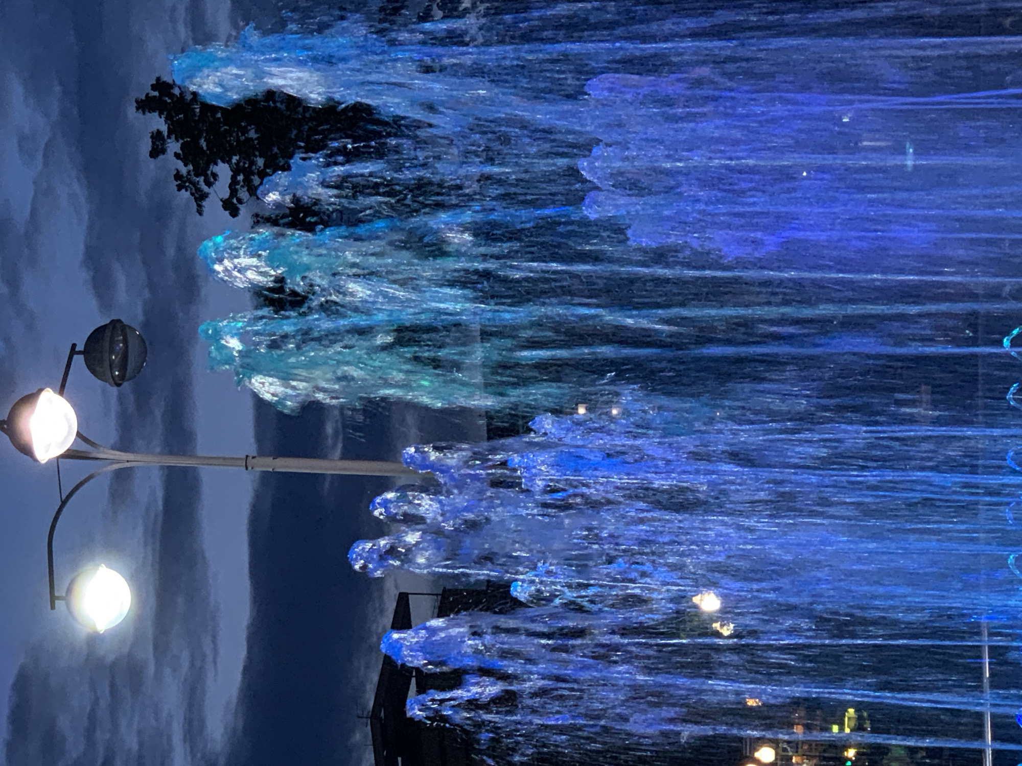 A night-time, eye-level, medium shot shows a fountain with illuminated blue water jets in front of a dimly lit, dark blue, cloudy sky. A lamp post with two globes stands to the left of center in the background. The top globe is illuminated white and is closer to the viewer. The bottom globe is unlit and appears dark. To the right of the lamp post, a dark silhouetted tree is visible. In the far background, a dark building with a balcony is partially visible. Several water jets, lit in various shades of blue and teal, shoot upwards and outwards from the bottom of the frame. The water is captured in motion, appearing frothy and dynamic. The scene is dark, with the primary light source coming from the illuminated fountain and the street lamp.