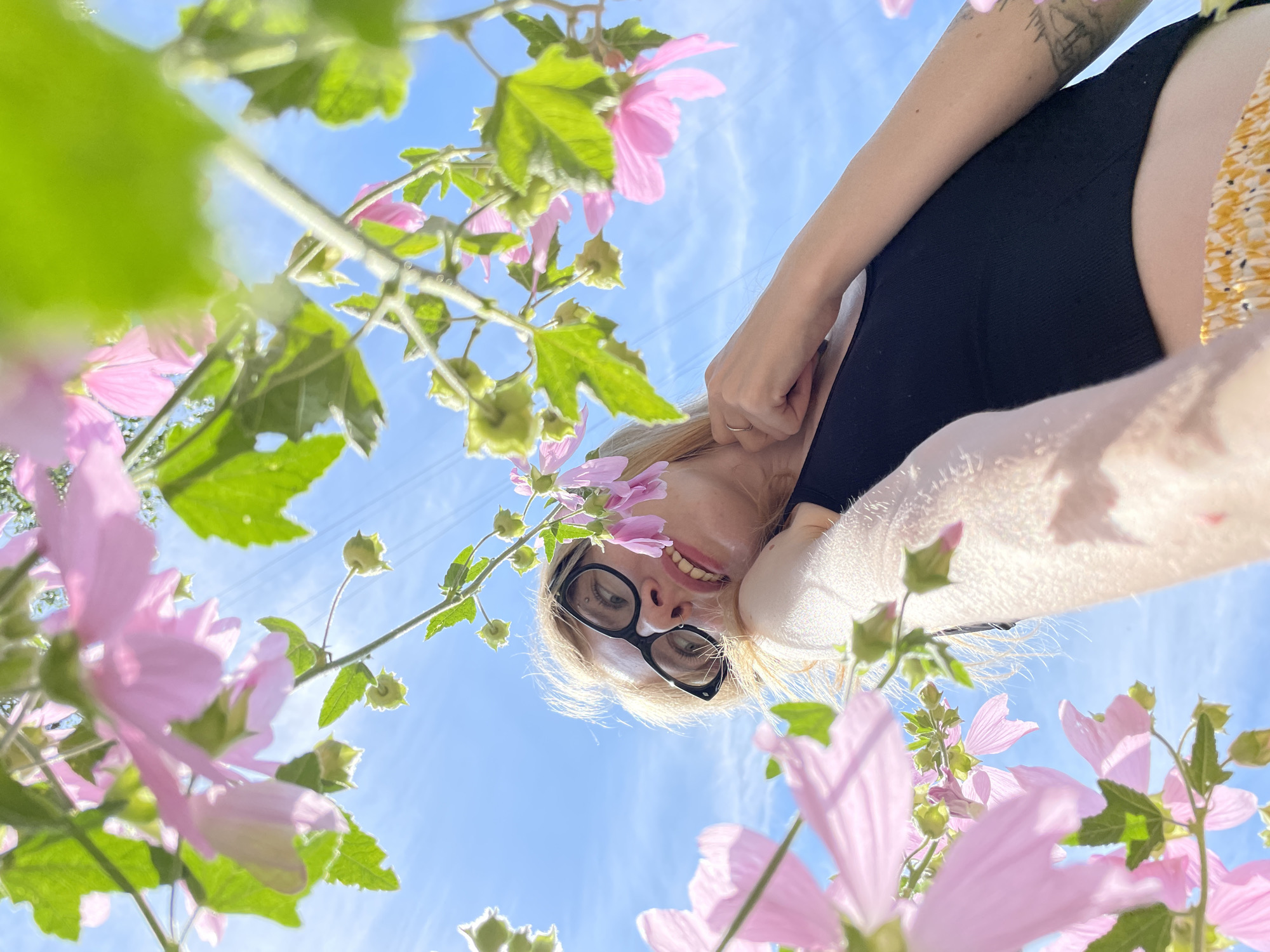 A low-angle shot shows a woman with blonde hair and glasses looking up at the camera, framed by pink flowers and green leaves against a bright blue sky. She is wearing a black tank top and a yellow and white patterned skirt. Her left hand is touching her chin, and her right arm is resting on her chest. The scene appears to be outdoors, possibly in a park or garden, with power lines visible in the distance. The sunlight creates lens flare and highlights on the flowers and the woman's skin. There is no discernible information in the image to indicate the location is Izhevsk, Russia.