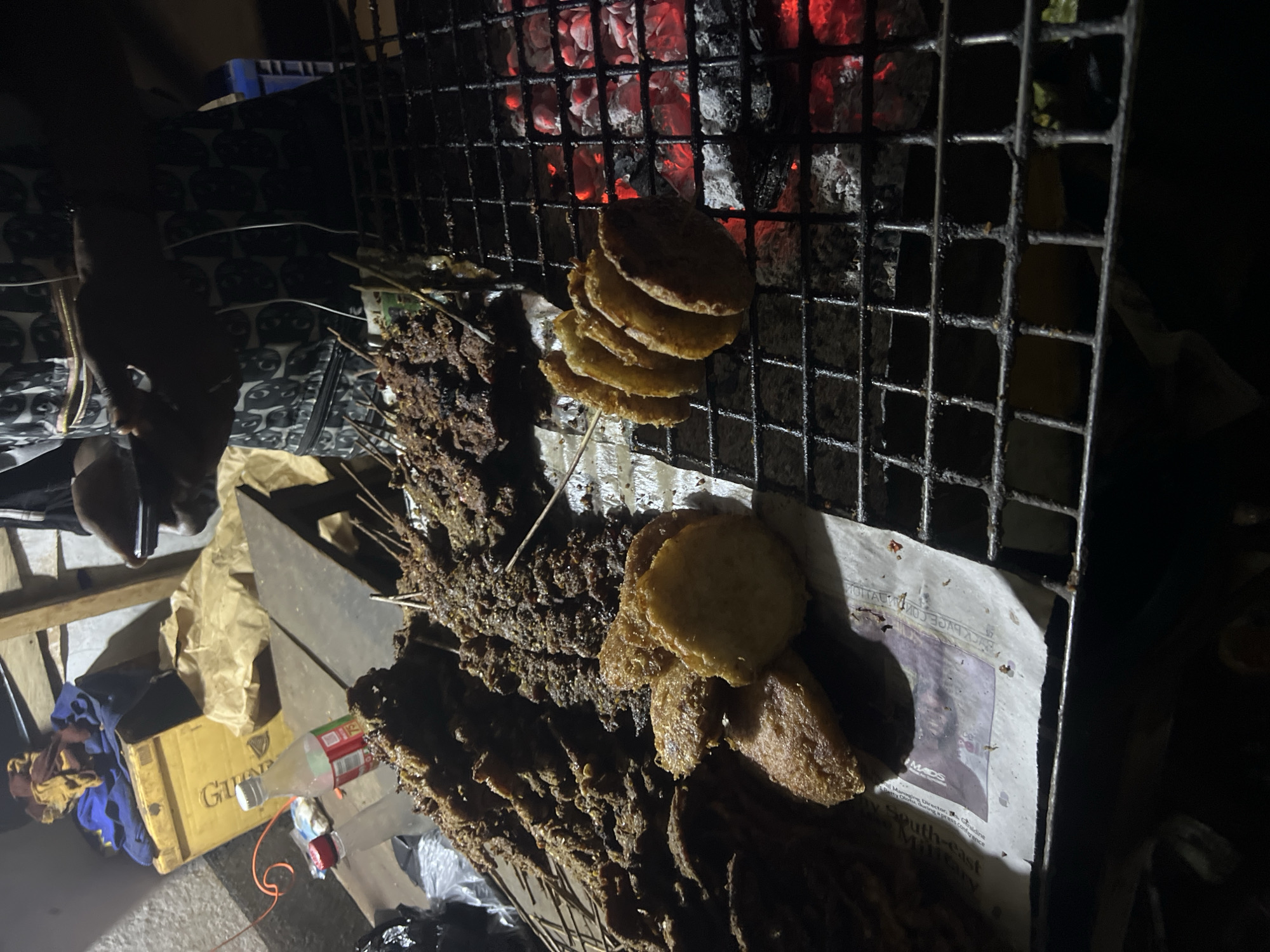 In Wumat, Nigeria, a street vendor grills meat skewers and fried plantains over glowing coals. The food is laid out on a metal grate, with some pieces stacked high on skewers and others as flat, golden-brown discs. A bottle of drink sits nearby, alongside what appears to be a yellow cooler filled with fabric. The scene is dimly lit, suggesting it is nighttime, and the vendor, partially visible, is holding a mobile phone. The environment is rustic, with a wooden structure in the background and discarded paper lining the serving area.