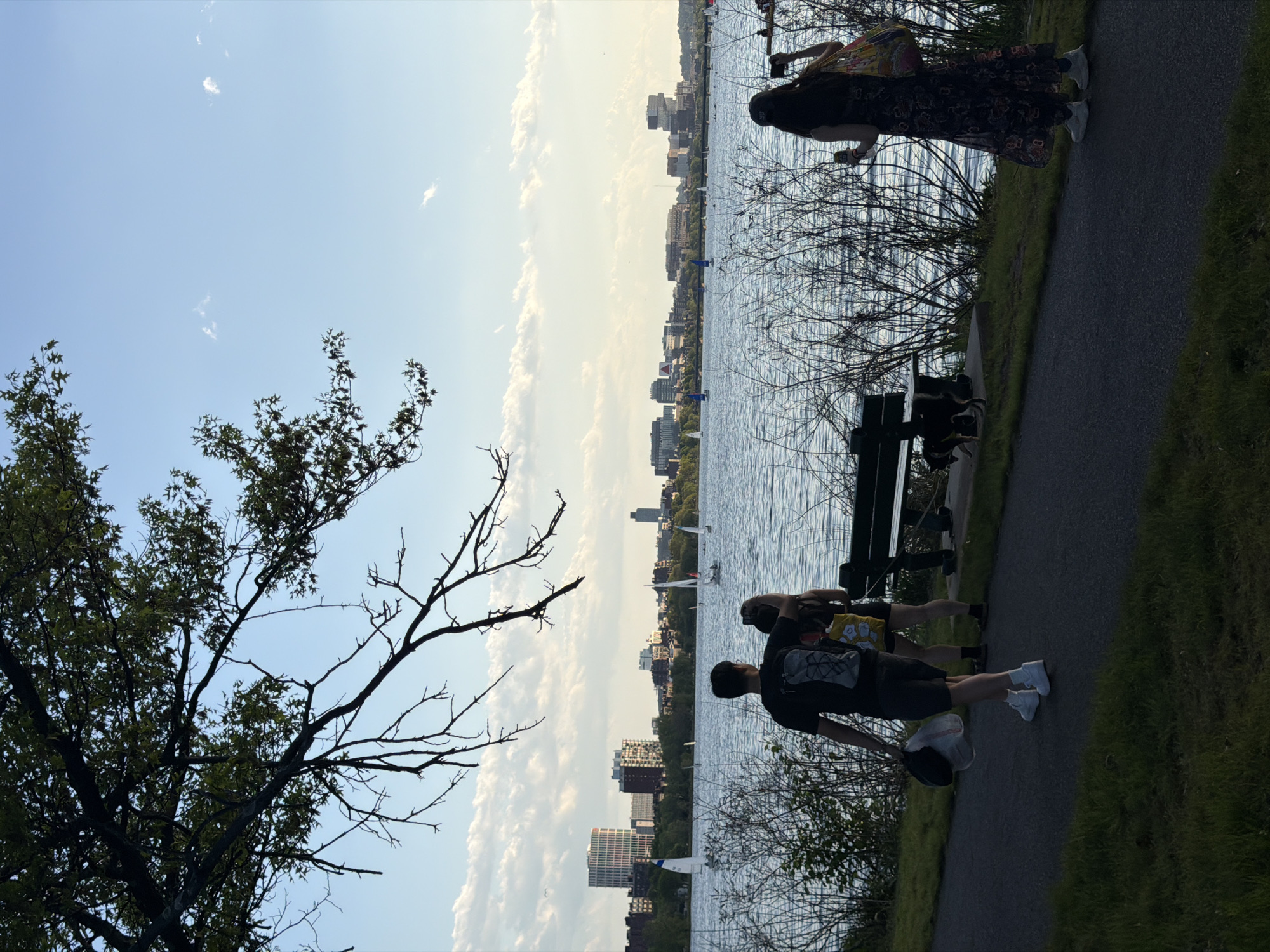 The image depicts a scene in Boston, Massachusetts, near the Charles River. The view includes the river, buildings in the city skyline, trees, and a walkway. There are people present on the walkway: one individual stands near the edge of the path looking out at the water and two people are sitting on a park bench. Several sailboats are on the water. The sky is blue with scattered clouds.