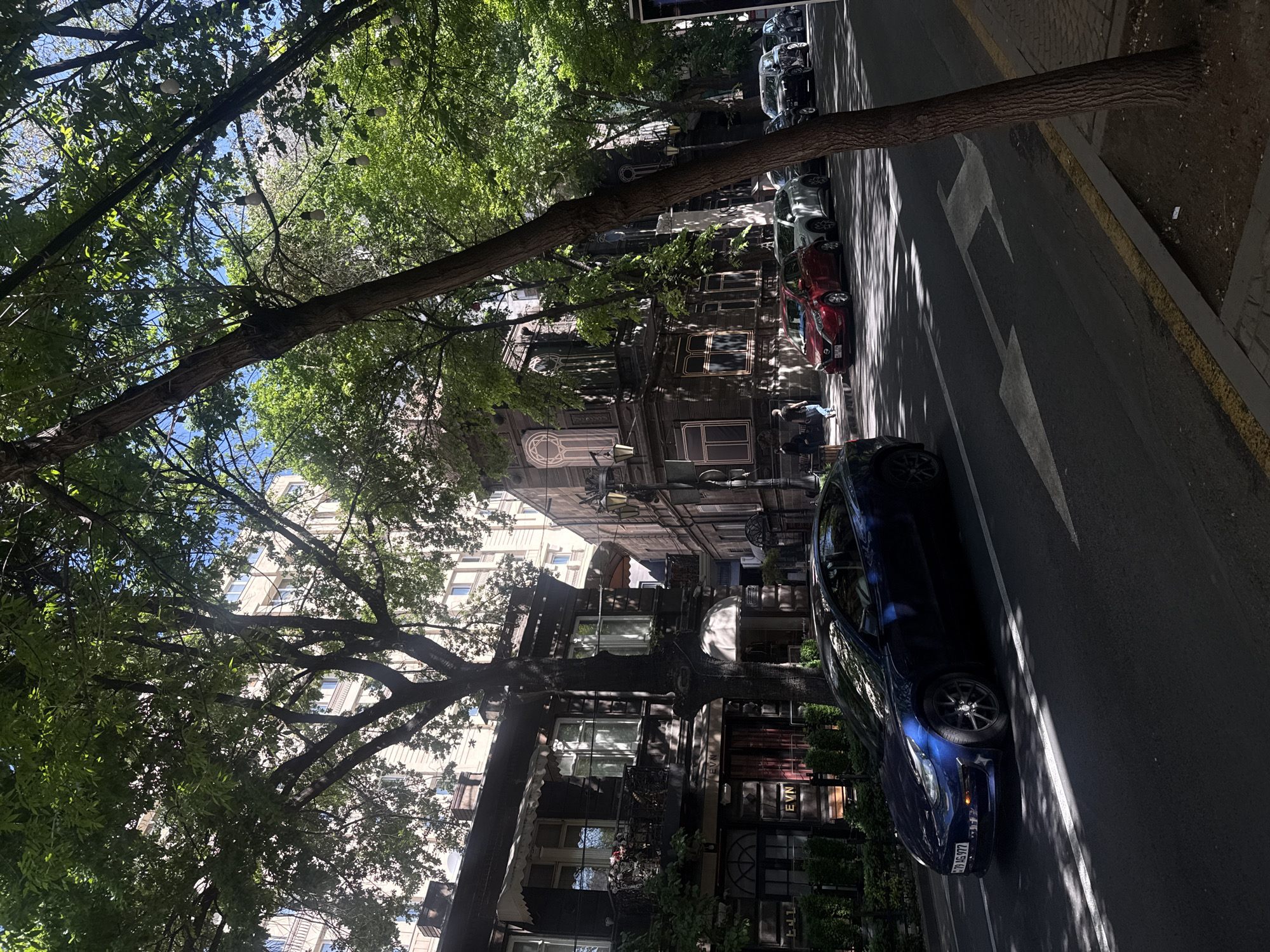 The image shows a street scene in Yerevan, Armenia. The view is of a tree-lined street with buildings lining the left side. A dark blue car is parked on the right side of the street. In the background are parked cars and pedestrians. The light is dappled due to the trees overhead, creating shadows on the road. The street has painted arrow markings.