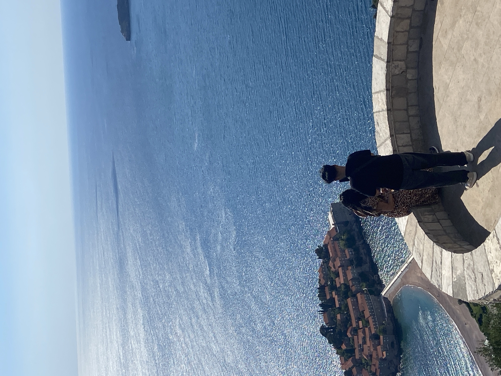 Two individuals are captured in an outdoor, daytime scene, standing on a curved stone viewing platform or balcony. The image, originally rotated, shows them overlooking a vast expanse of sparkling blue water, which is the Adriatic Sea.
In the foreground, a male figure is visible from the waist up, wearing a dark long-sleeved top, dark trousers, and light-colored shoes. He is positioned with his back mostly to the camera, looking out towards the scenic view. A second person, a female, is partially visible in front of him, wearing a leopard-print top or dress, also looking out. The paved ground of the platform is light-colored, and a stone railing separates the platform from the sea. Distinct shadows on the pavement indicate bright sunshine.
Below and to the right of the platform, the iconic island-town of Sveti Stefan in Montenegro is clearly visible. It features numerous compact, terracotta-roofed buildings clustered together on a small landmass connected to the mainland by a narrow causeway. A sandy beach with calm turquoise water is also apparent near the island. The sea glistens with strong sun reflections, particularly in the middle distance. The sky above is a clear, light blue, meeting the horizon line. No discernible text is visible in the media file.
